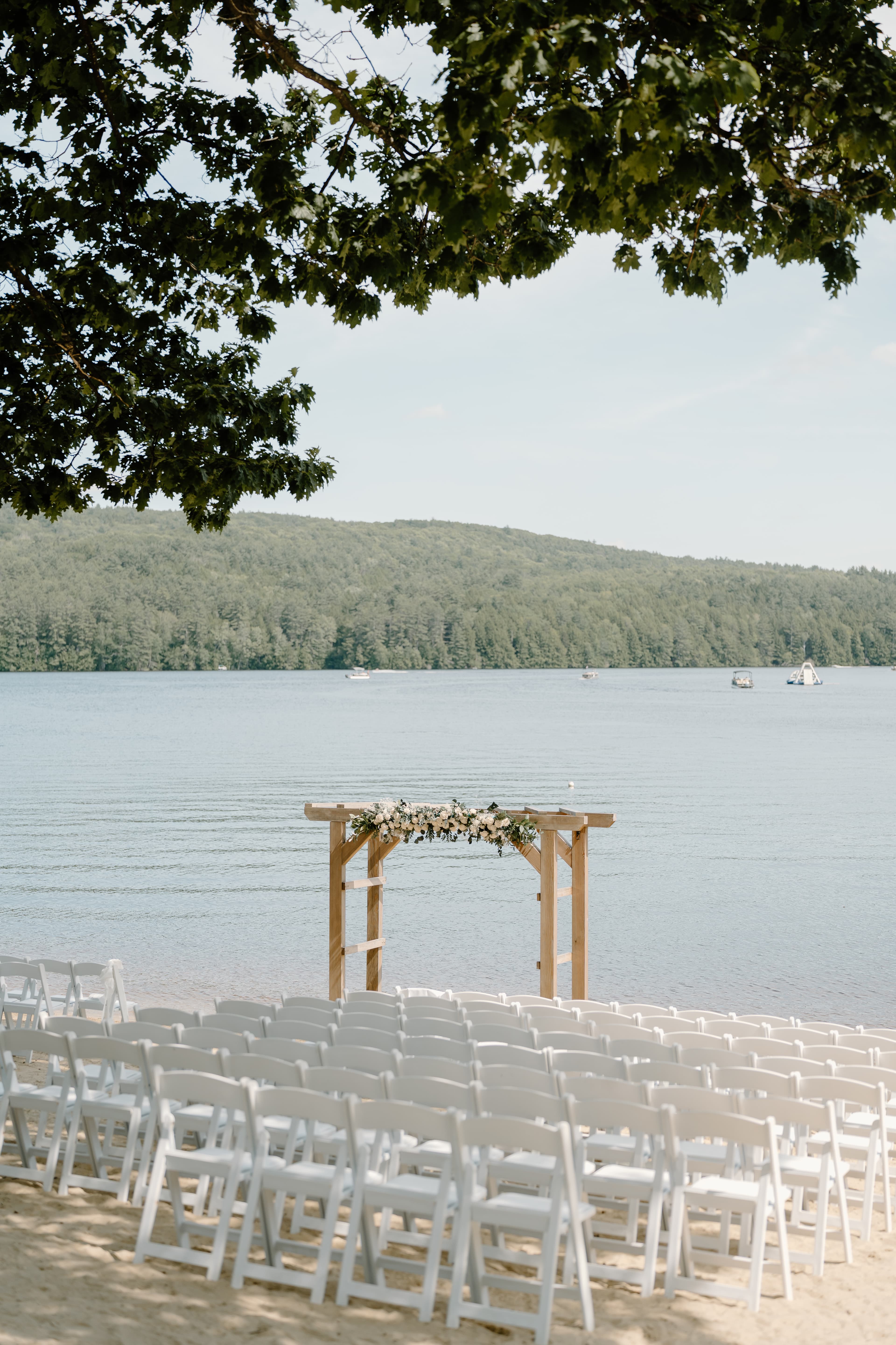 A scenic lakeside wedding setup featuring rows of white chairs and a floral arch.