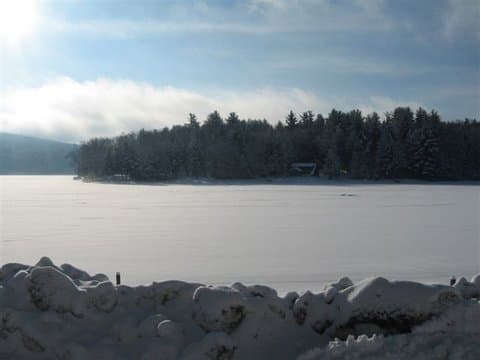 A snowy landscape featuring a frozen lake and tree-covered shoreline under a partly cloudy sky.