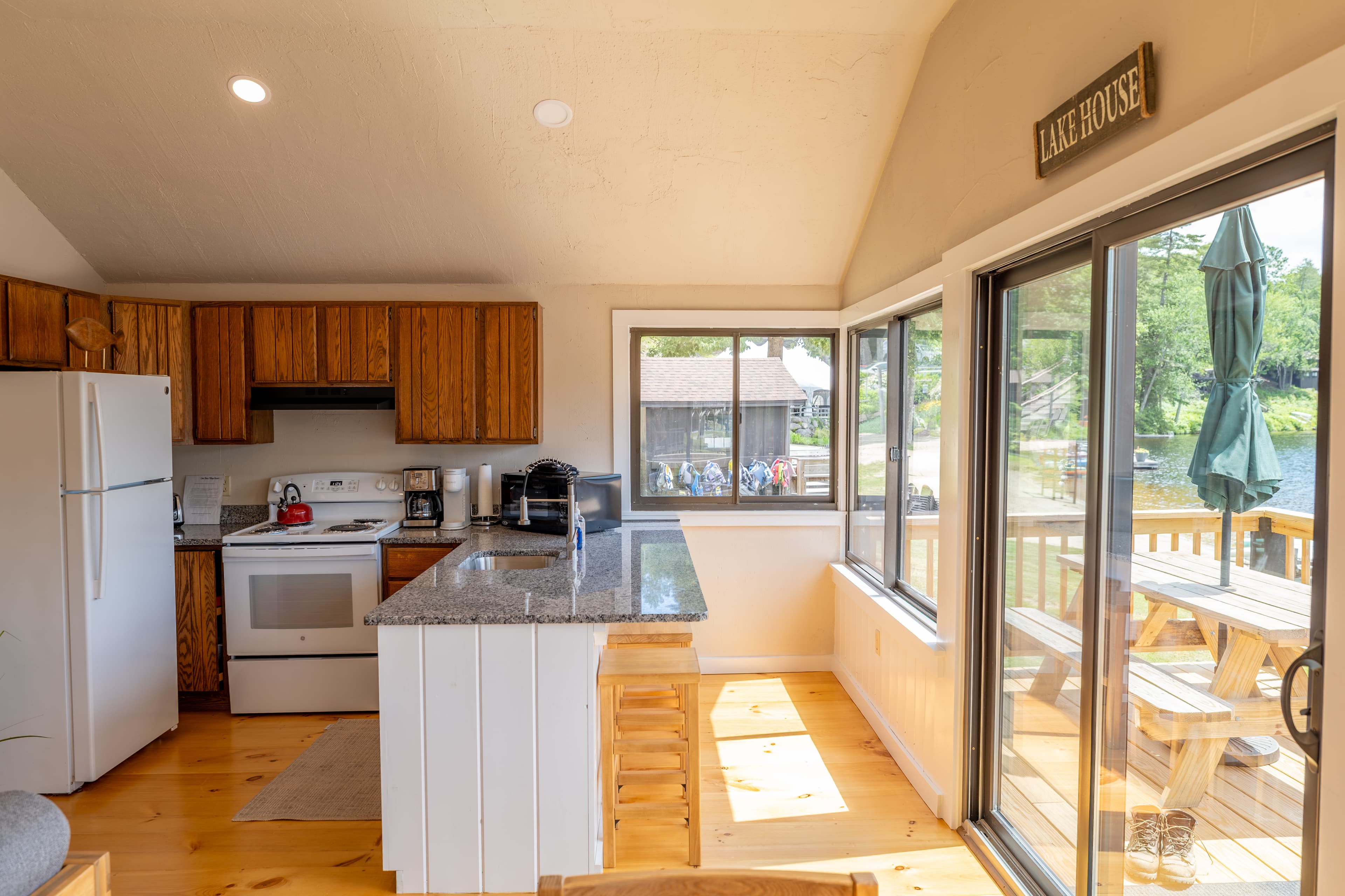 A bright cabin kitchen and dining area with a vaulted ceiling, wood cabinets, a white fridge, and a dining nook next to large windows.