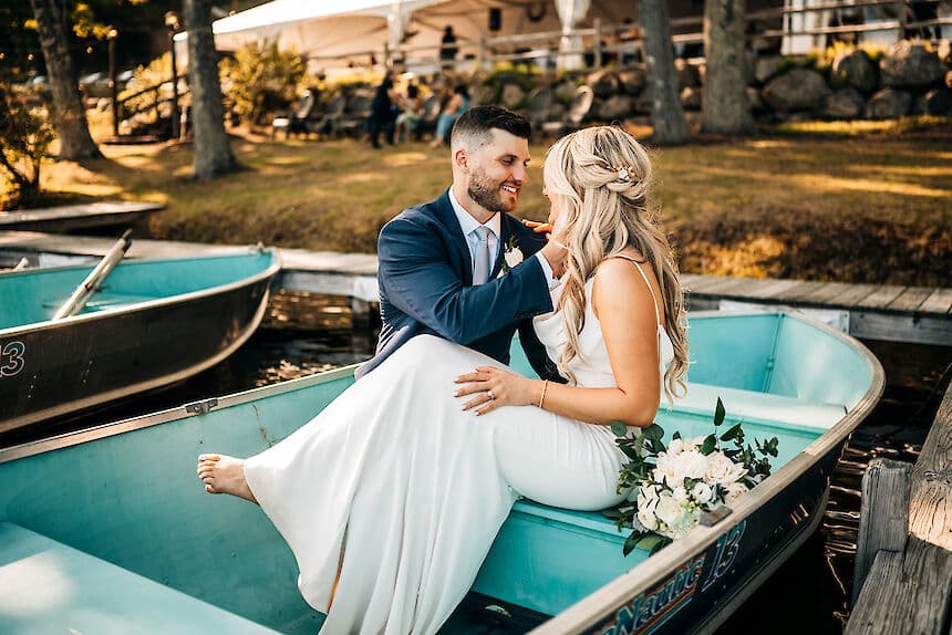 A smiling couple in formal attire sits on the edge of a small boat, surrounded by nature, as they share a tender moment.
