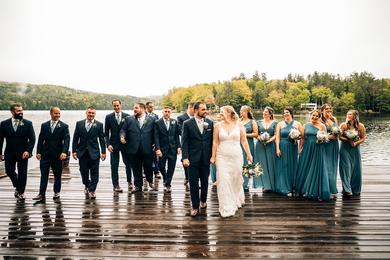 A bride and groom walk hand-in-hand with their wedding party on a dock by a lake.