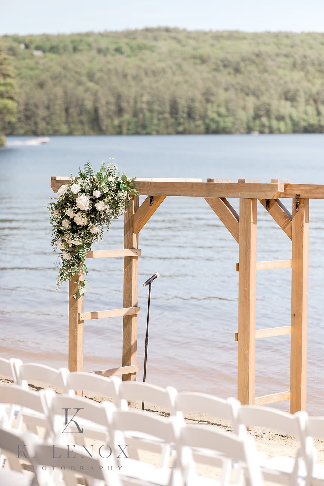 A wooden wedding arch adorned with flowers, set by a calm lake and white chairs in the foreground.