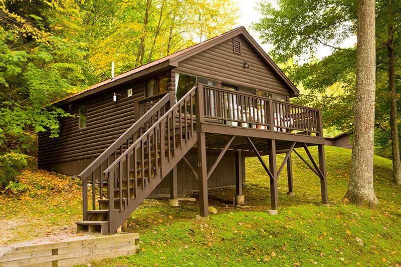 A brown wooden cabin with a deck, surrounded by trees and autumn foliage.