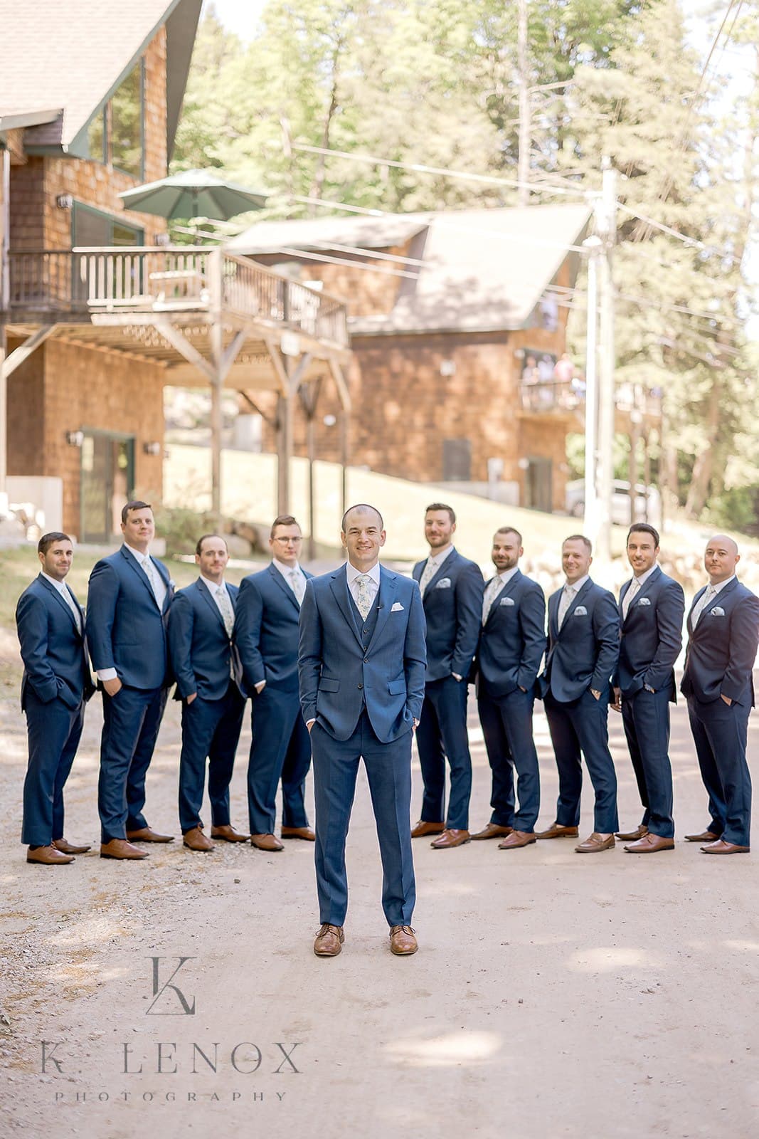 A groom stands confidently in front of his groomsmen on a dirt path flanked by wooden cabins.