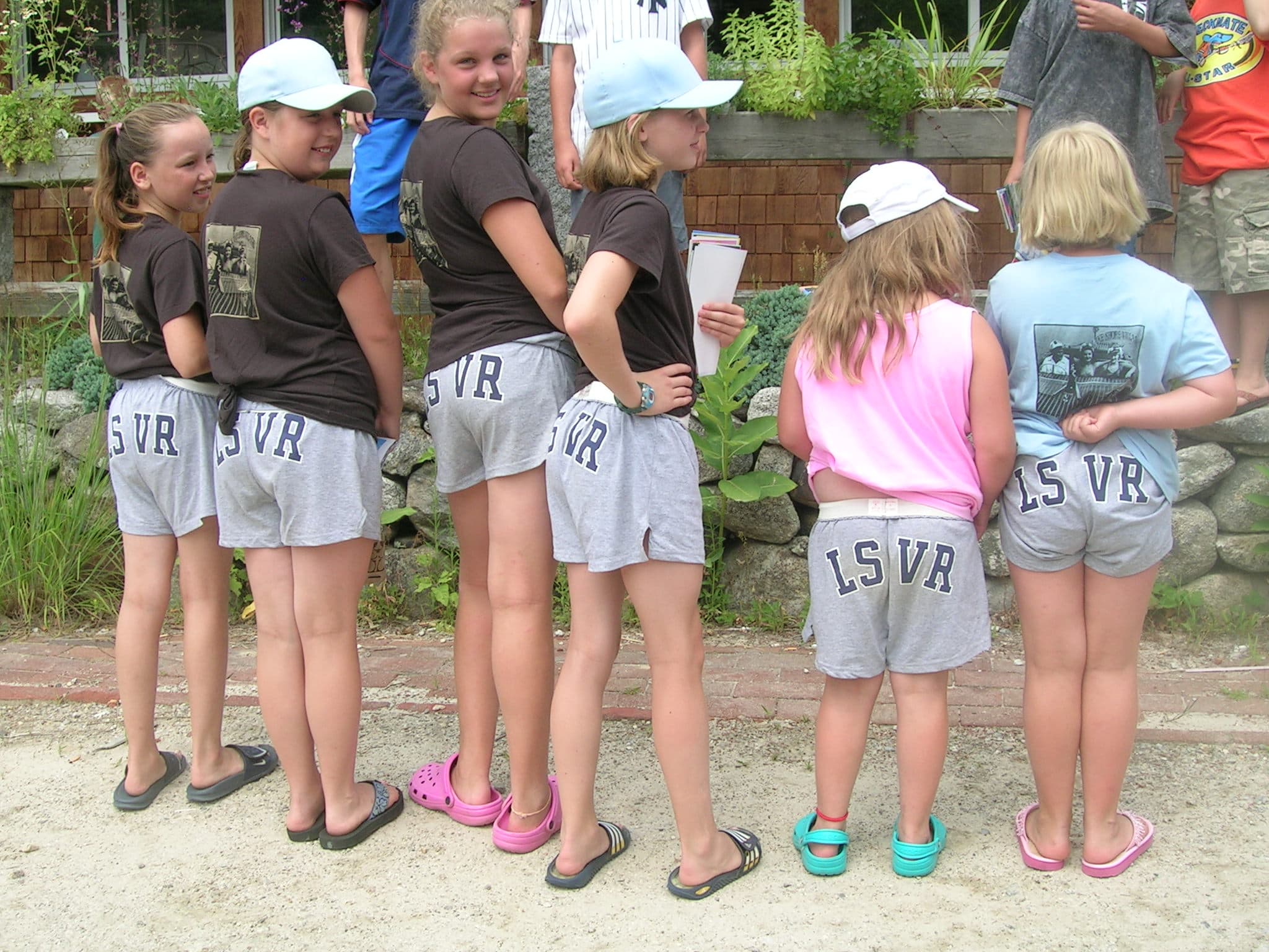 A group of children in matching gray shorts and t-shirts stands with their backs to the camera.