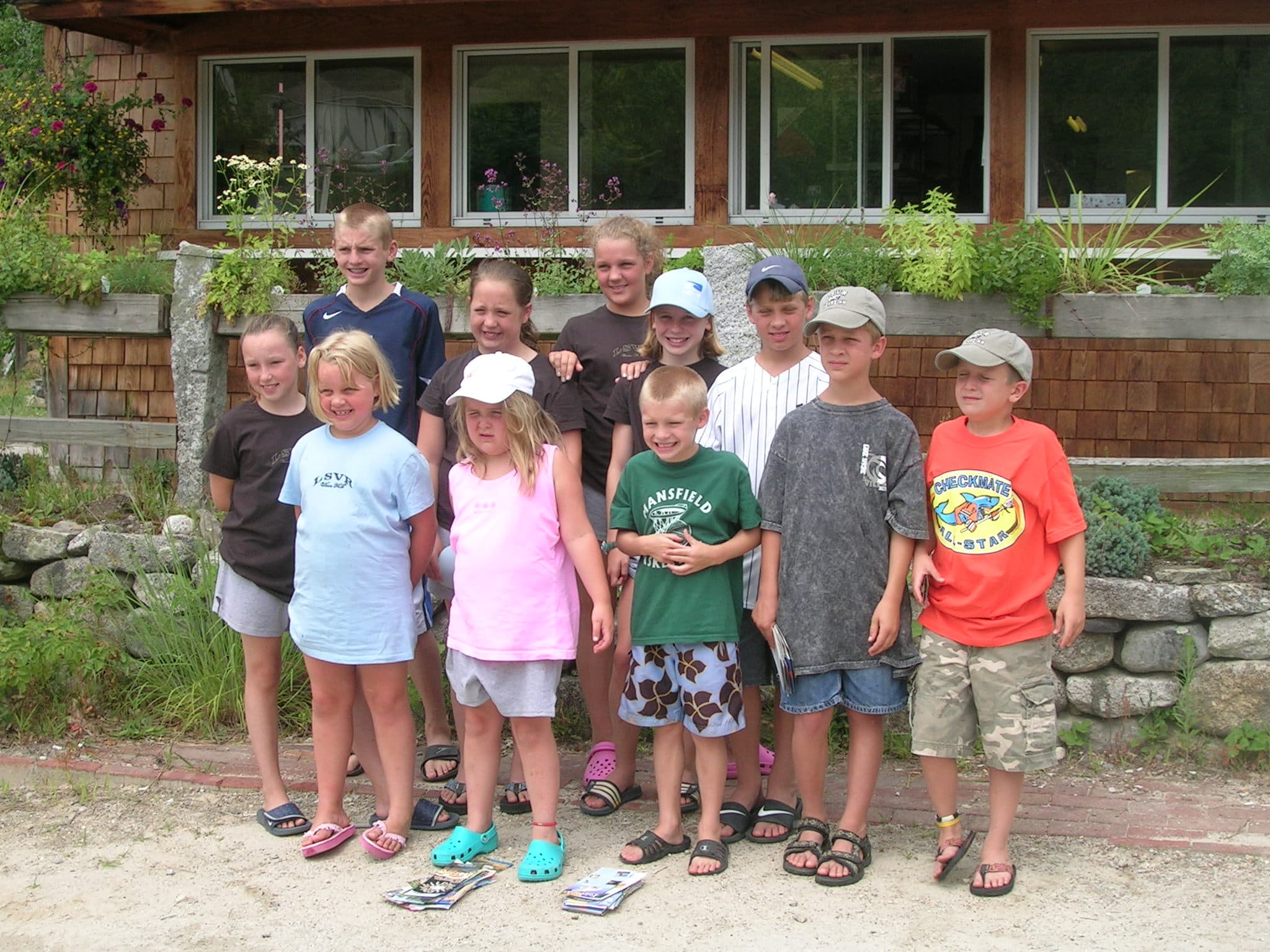 A group of ten children in casual summer clothing pose together outdoors in front of a rustic building with flowers.