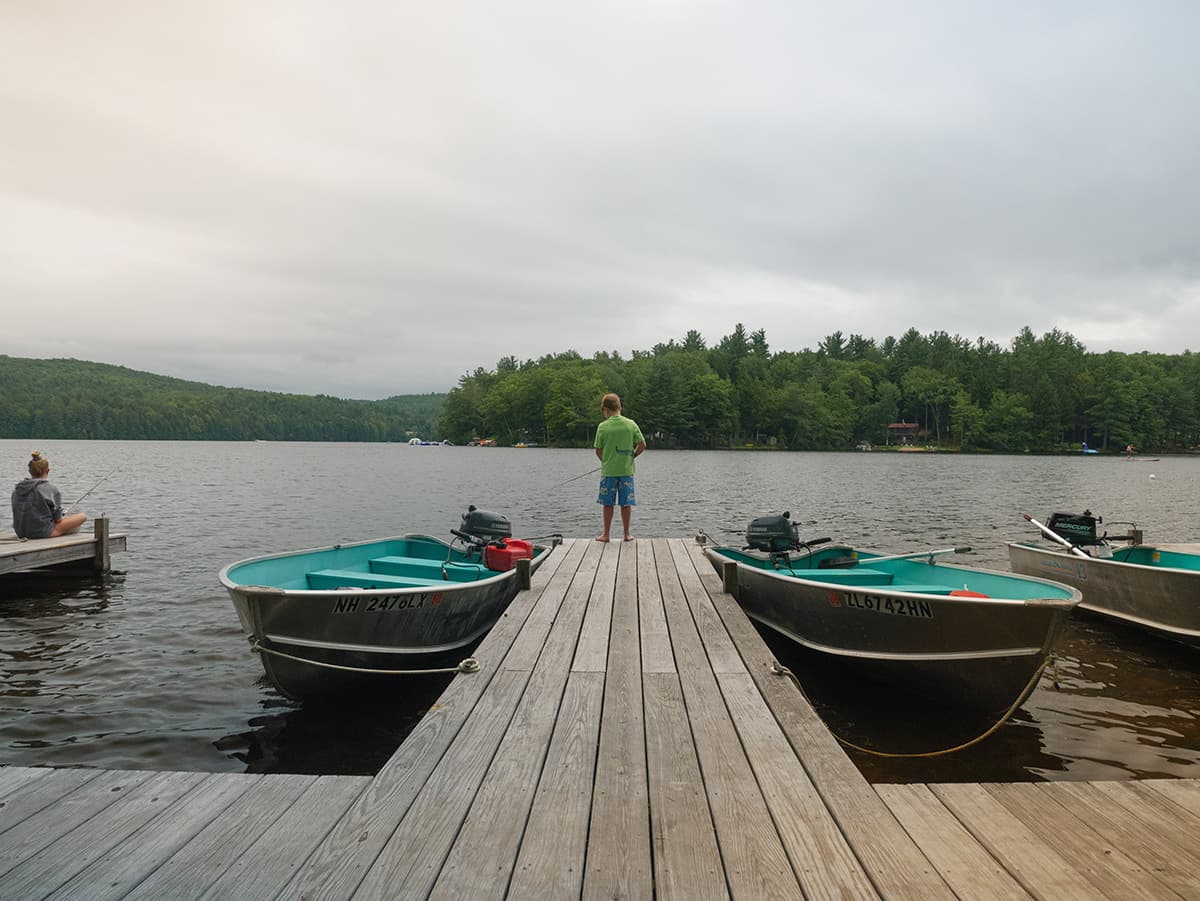 A person stands on a wooden dock facing a calm lake with boats nearby and trees in the background.
