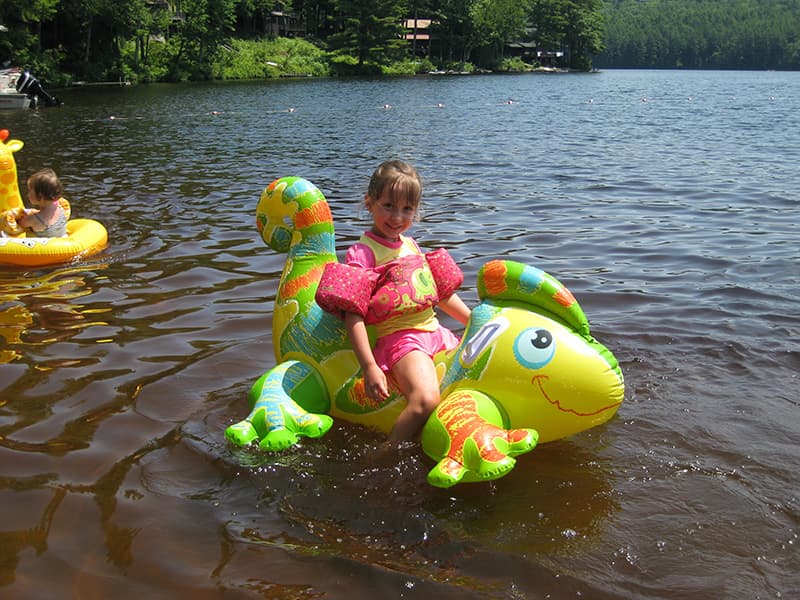 A young girl smiles while sitting on a colorful inflatable dinosaur in a lake, with another child in a smaller inflatable nearby.