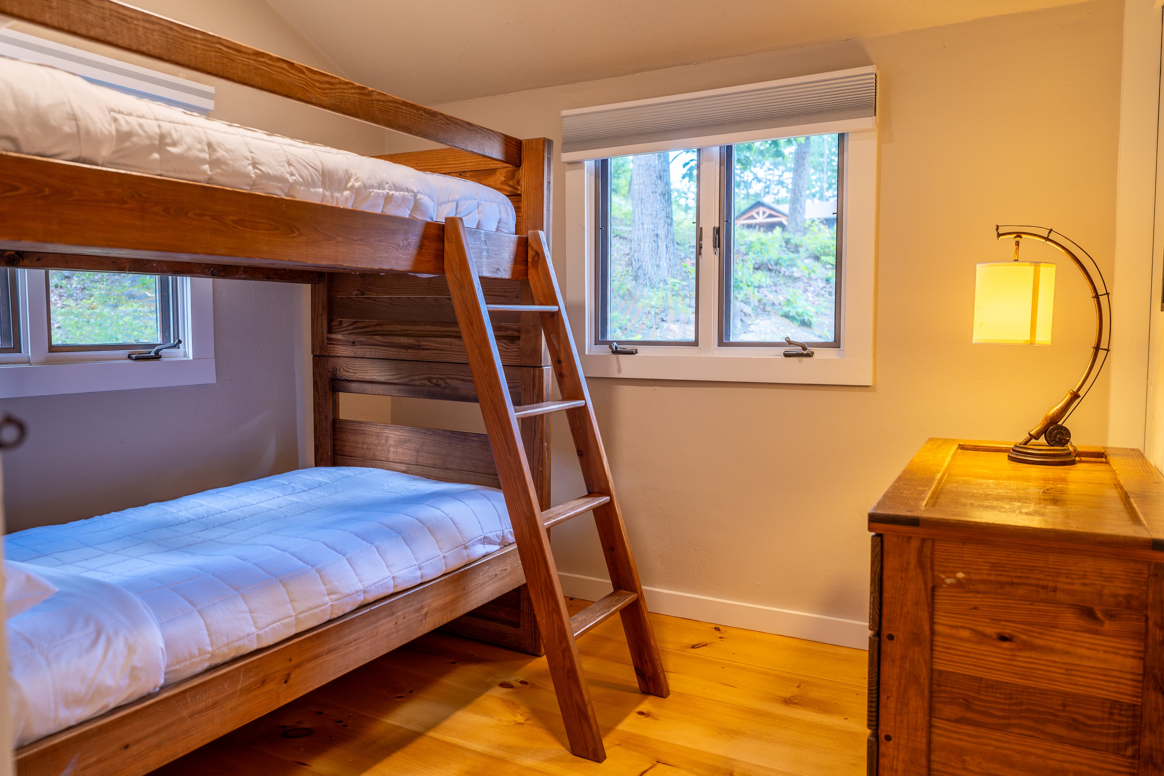 A simple cabin bedroom featuring a wooden bunk bed with a ladder, a small window with beige curtains, and a bedside lamp.
