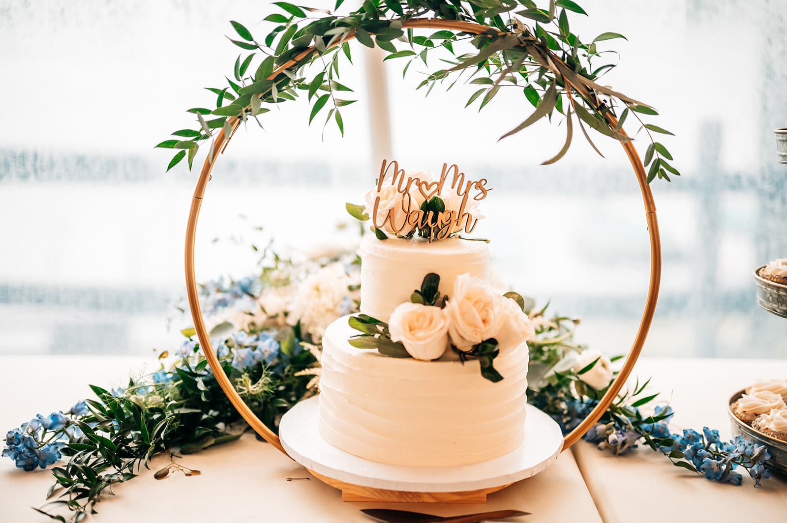 A two-tiered wedding cake decorated with white roses and greenery, topped with a wooden "Mr & Mrs Waugh" sign.