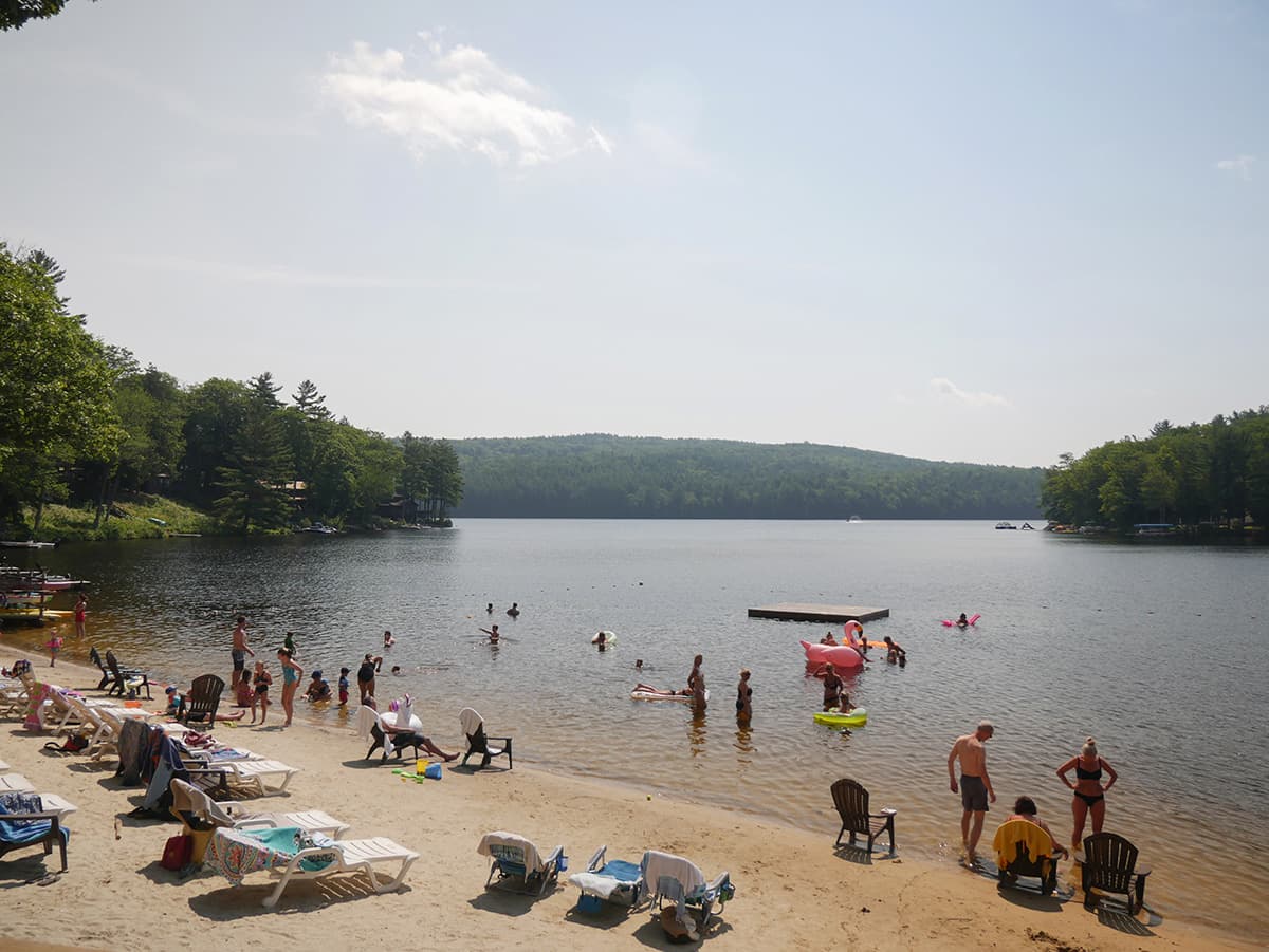 A sunny lakeside scene with people swimming and lounging on beach chairs.
