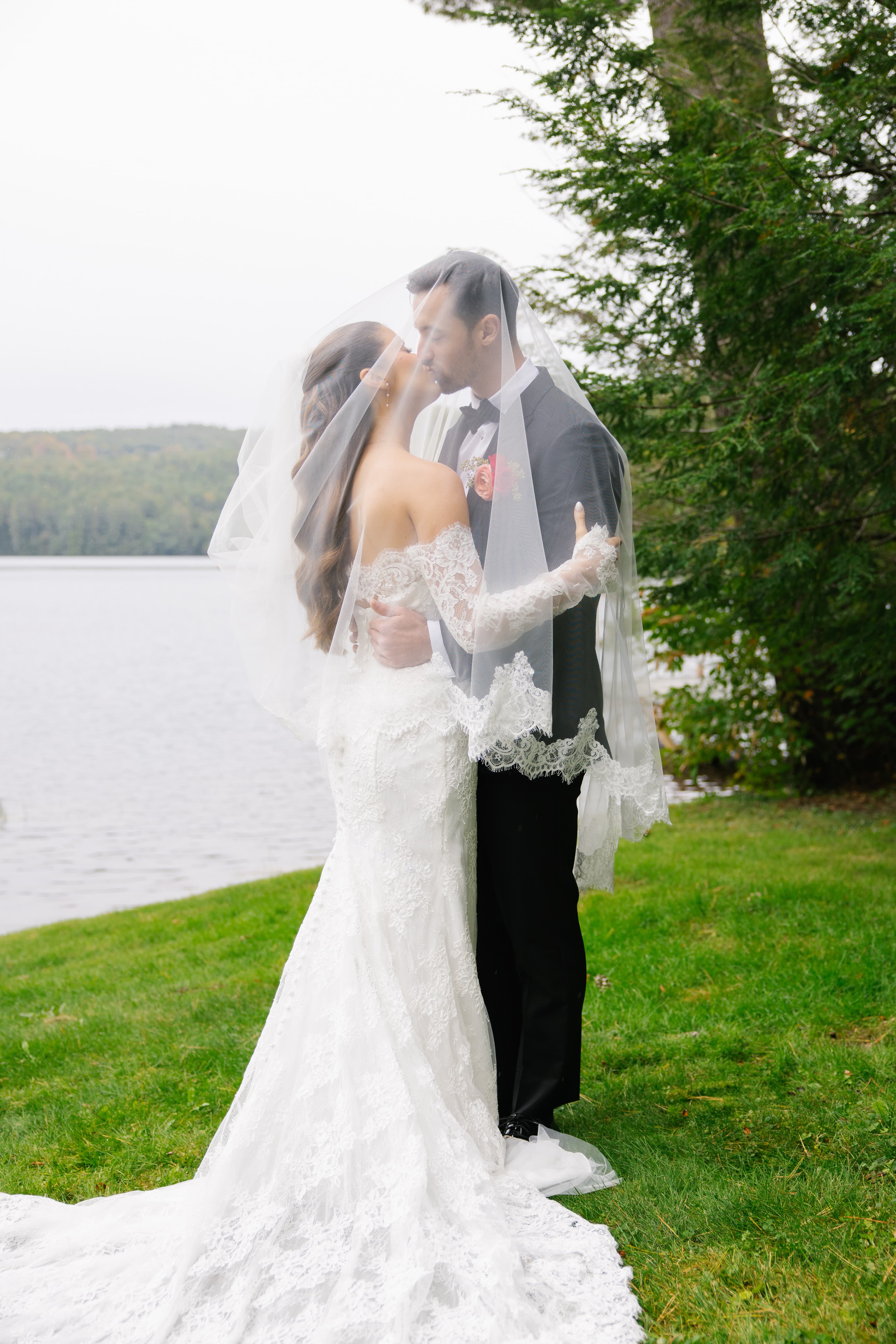 A couple shares a romantic kiss by a lake, surrounded by greenery and a soft veil.