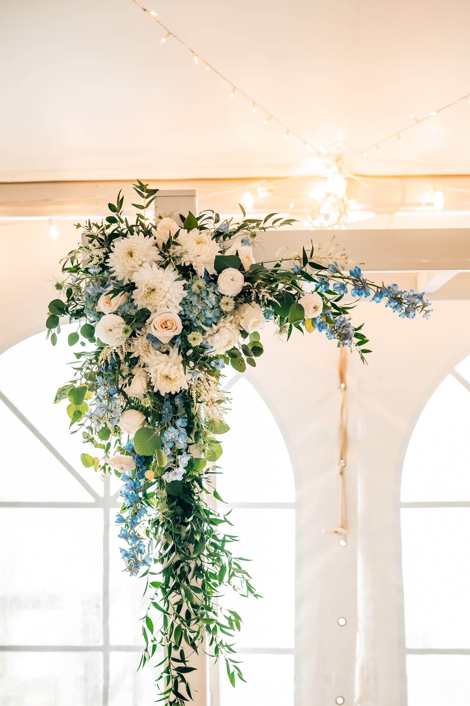 A floral arrangement with white and blue flowers hangs above a doorway in a well-lit tent.