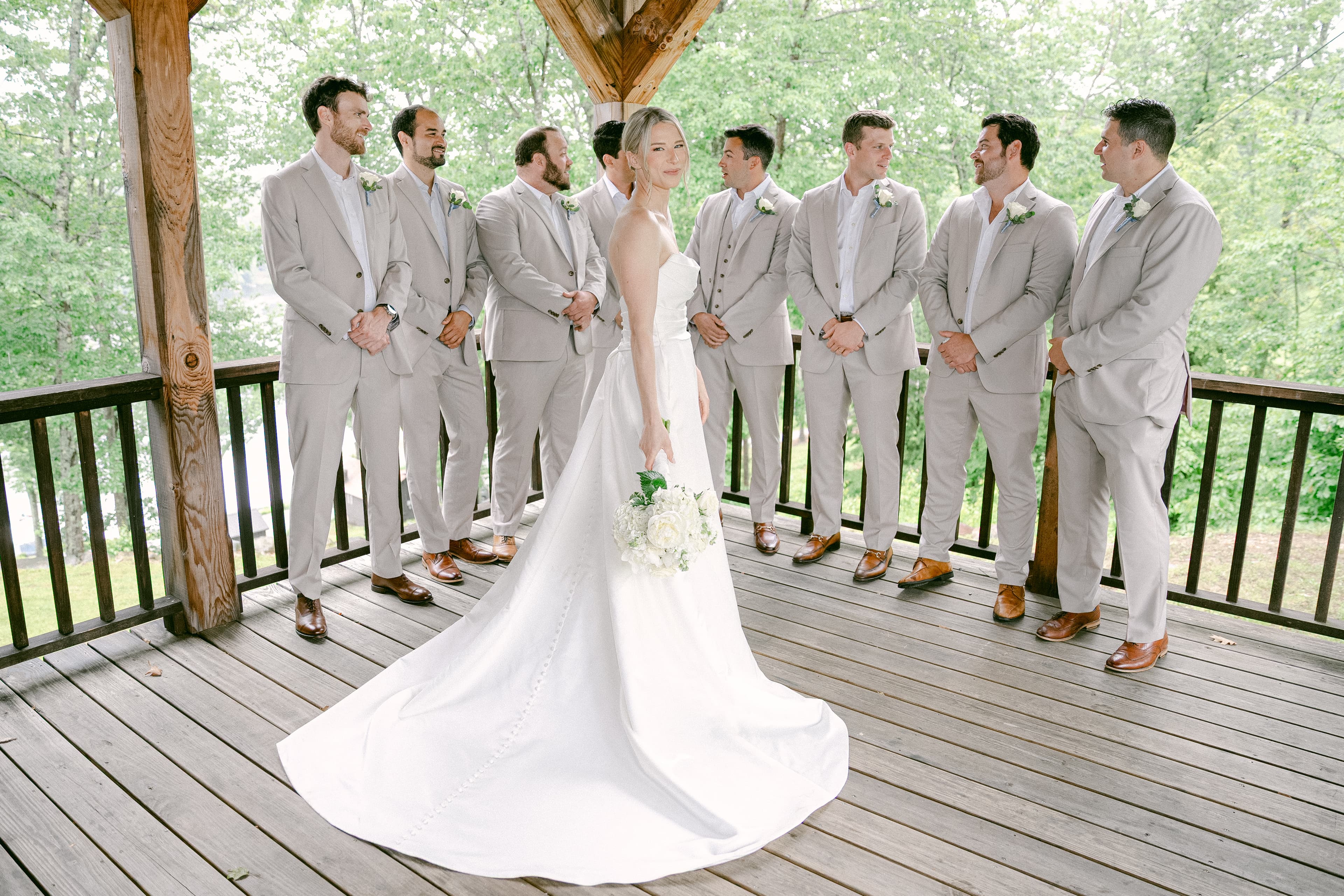 A bride in a white gown stands in front of a group of groomsmen in gray suits under a wooden gazebo.