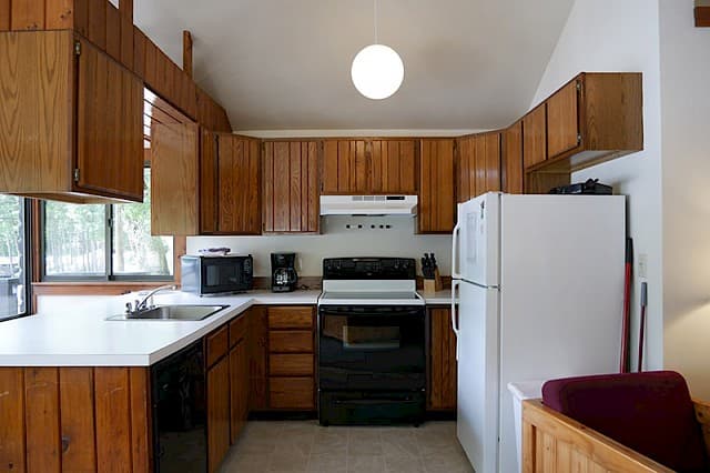 A cabin kitchen with a vaulted ceiling, wood paneling and cabinets, a white fridge, a black stove, and a single globe pendant light.