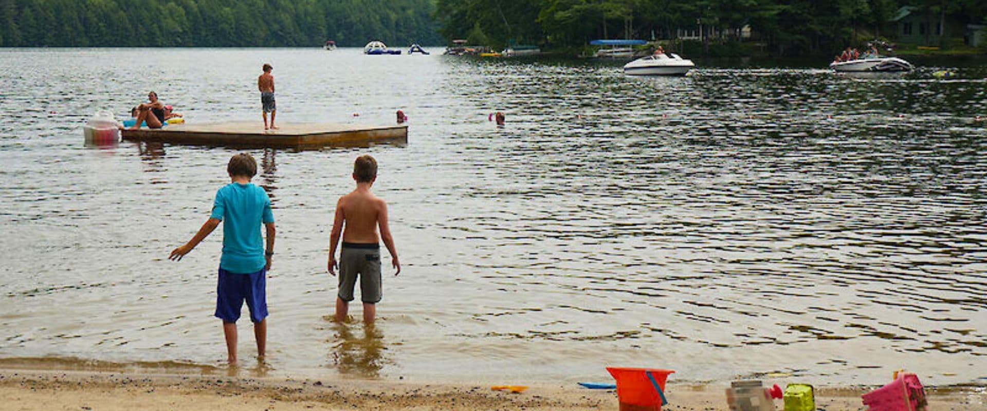 Two boys wade in a lake near a sandy beach, while others play on a dock and boats float in the background.
