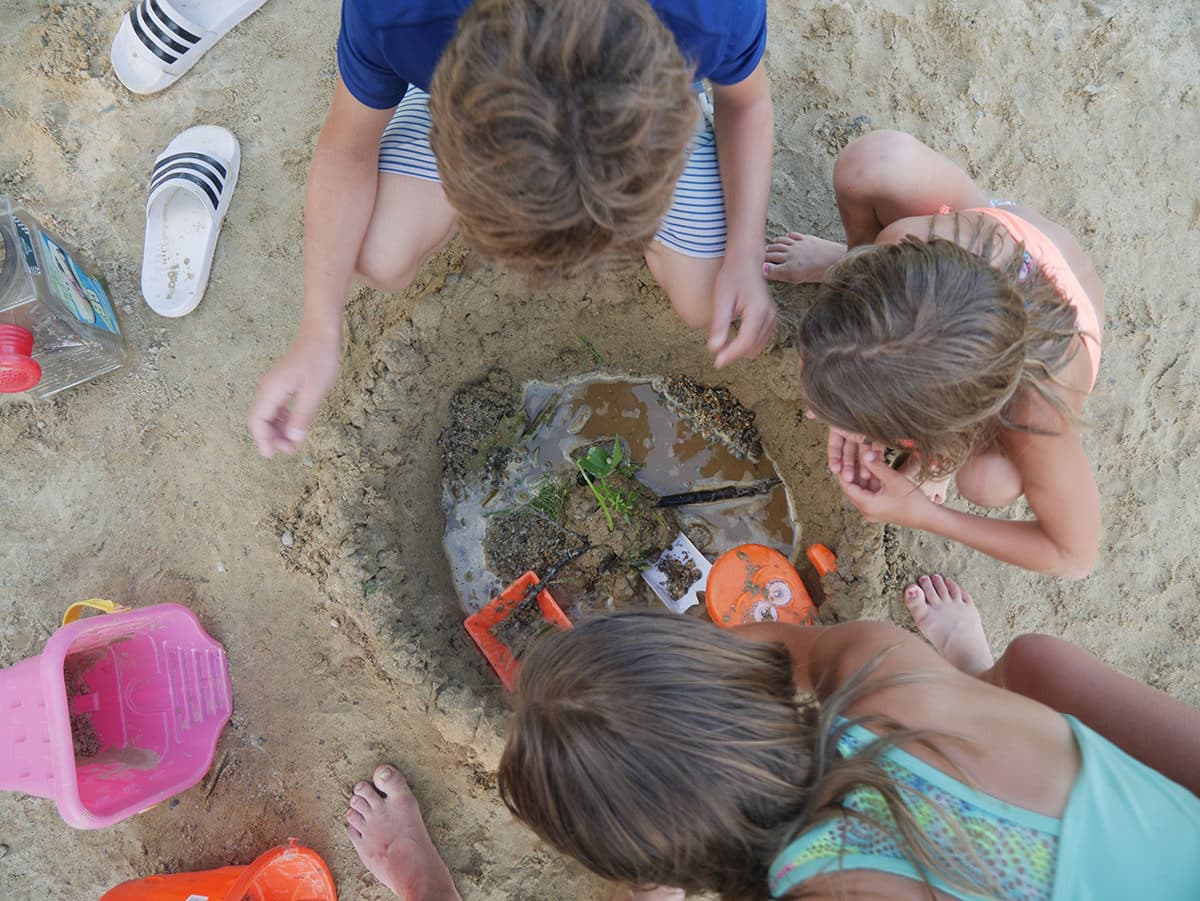 Children playing at the beach, exploring a sandy hole filled with water and toys.