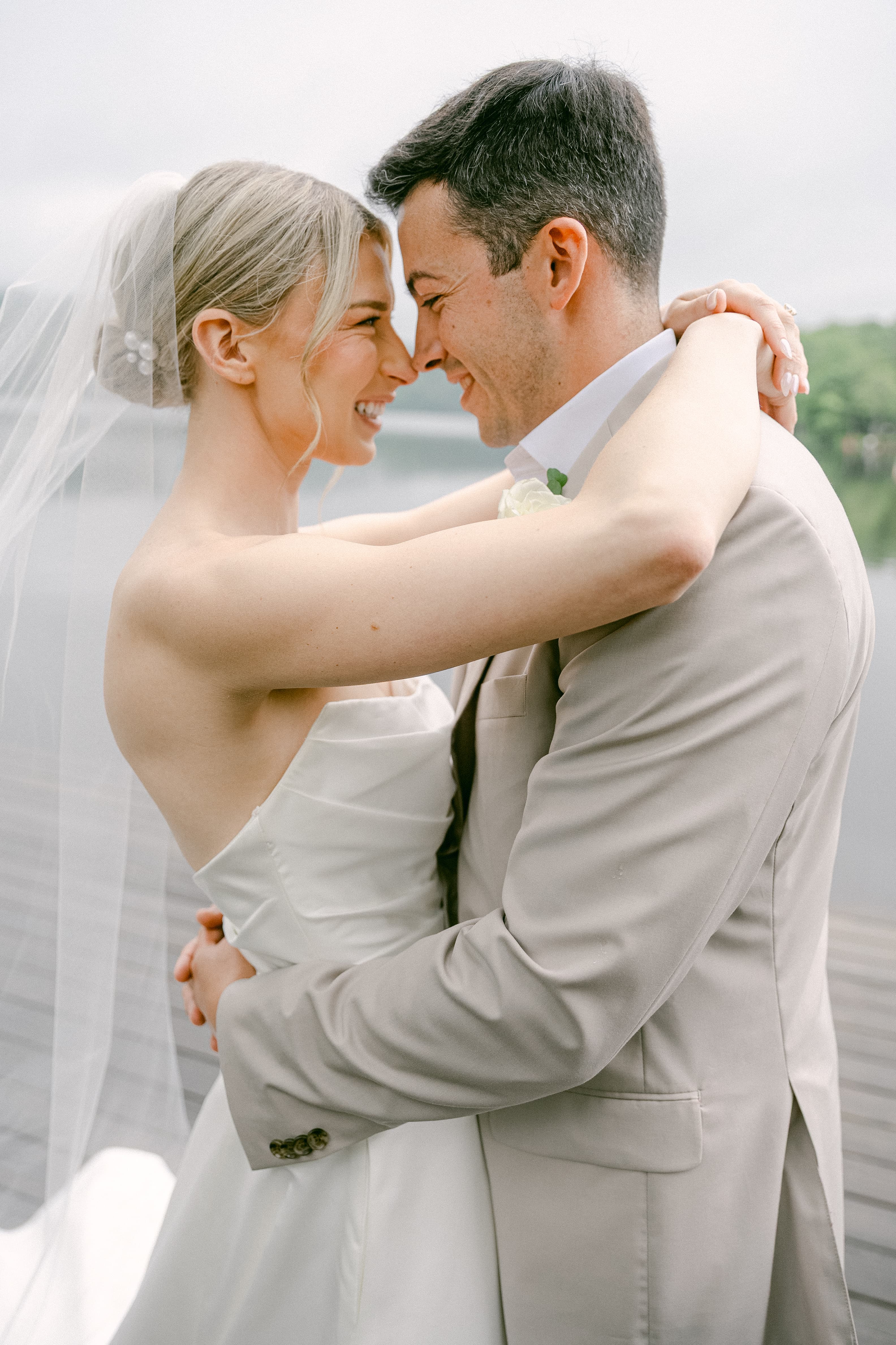 A bride and groom embrace joyfully by a serene lakeside.