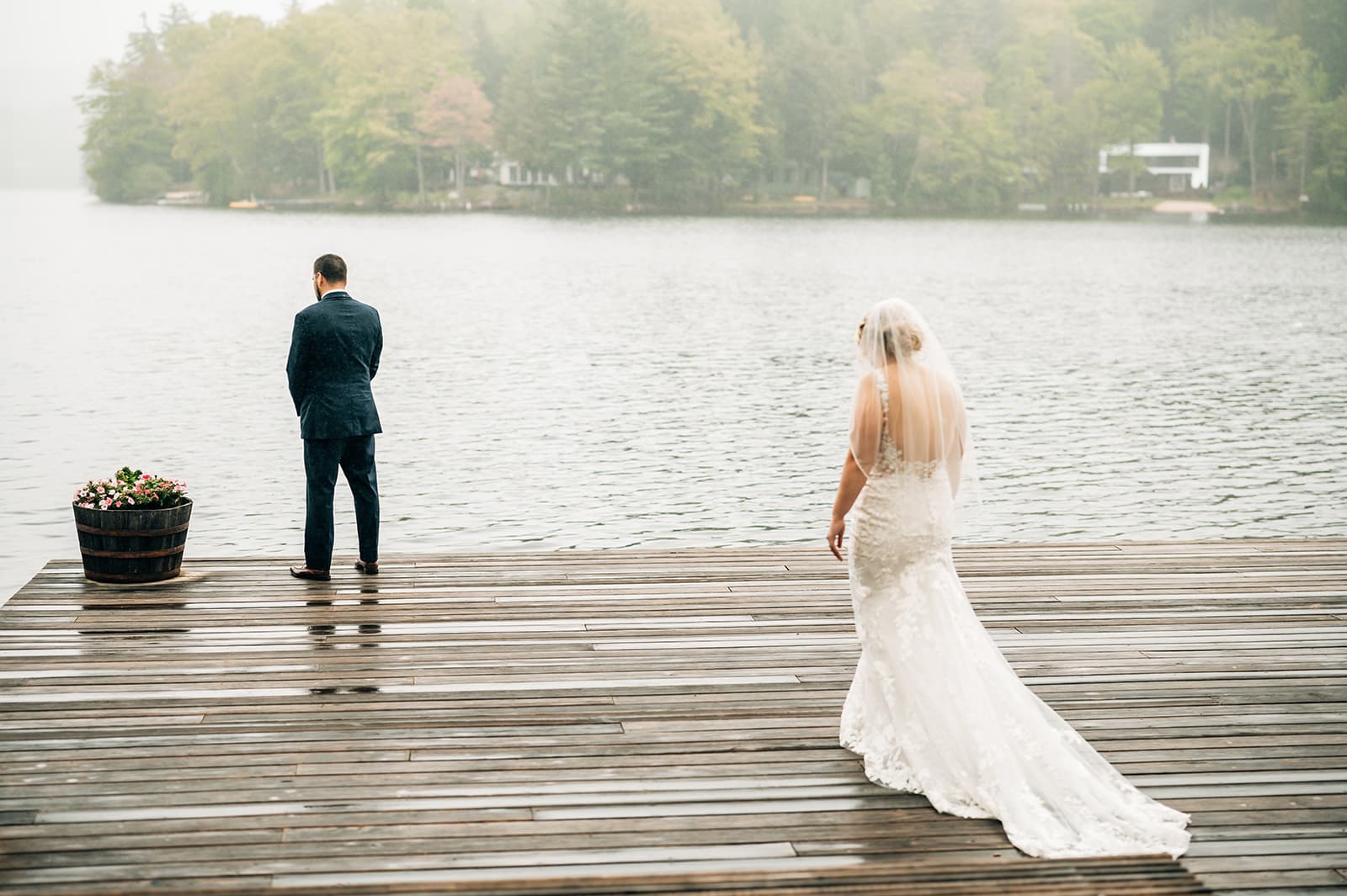 A bride approaches a groom standing on a dock by a misty lake.
