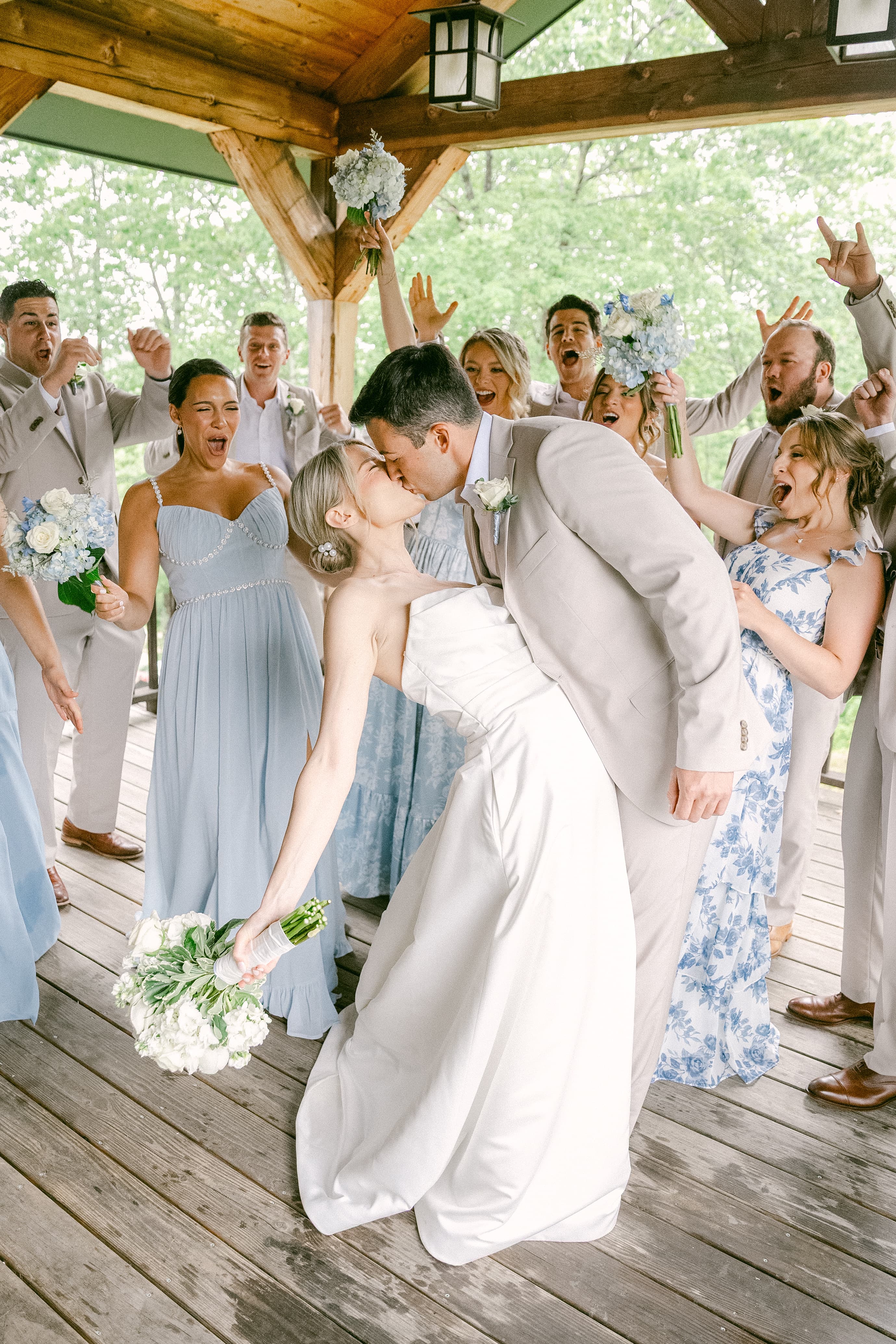 A bride and groom kiss joyfully while surrounded by excited wedding guests.