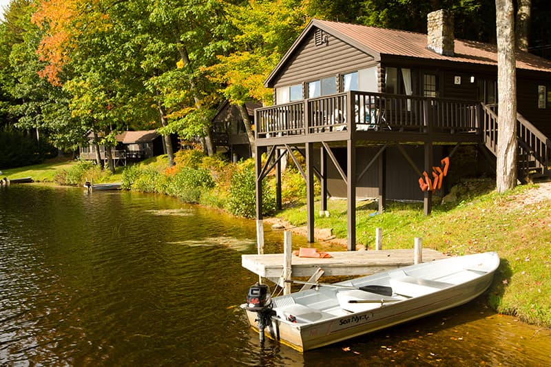 A wooden cabin with a deck overlooks a calm lake, with a small boat docked in front and vibrant autumn foliage surrounding the scene.