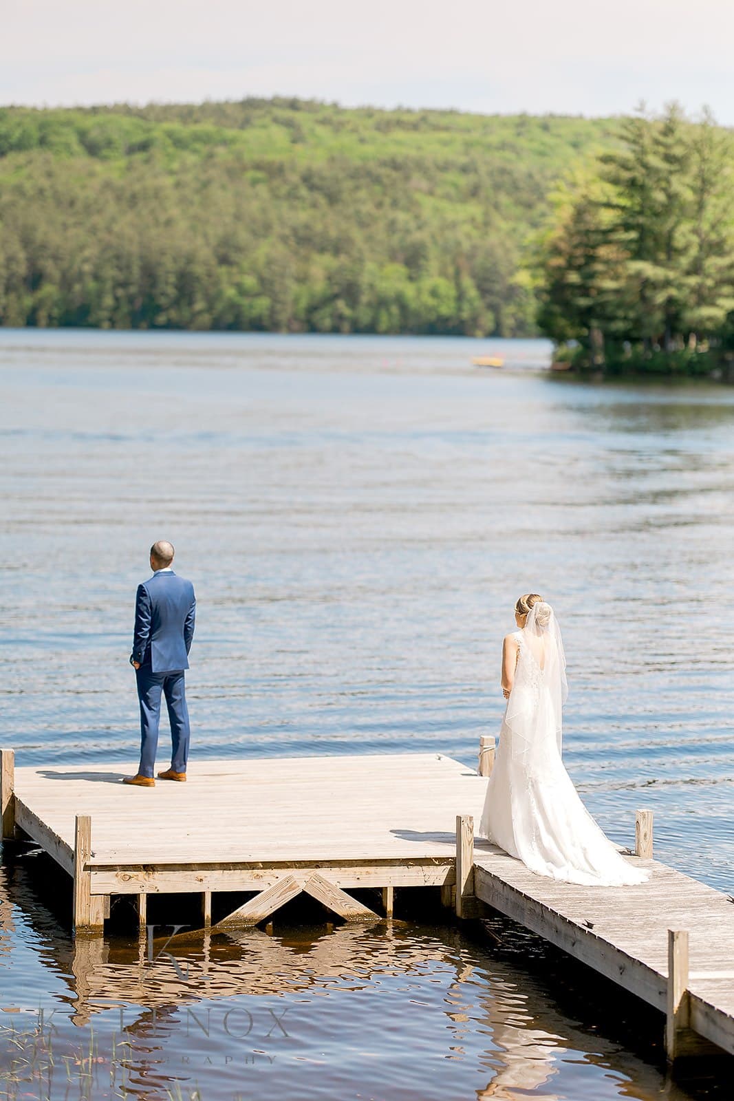A bride and groom stand on a dock overlooking a calm lake.