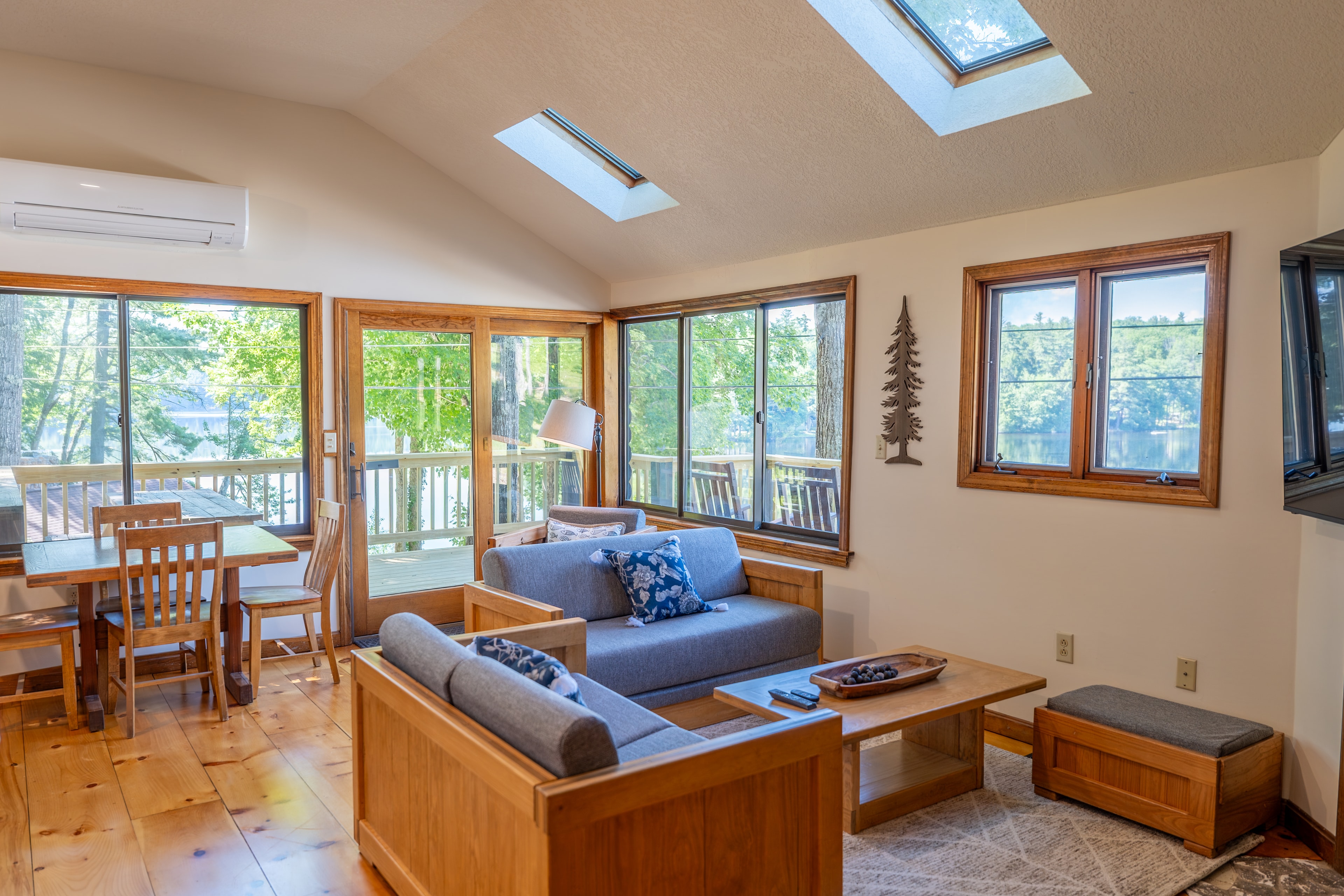 A bright cabin living area with a vaulted ceiling, skylights, a blue futon, a dining table, and large glass doors leading outdoors.