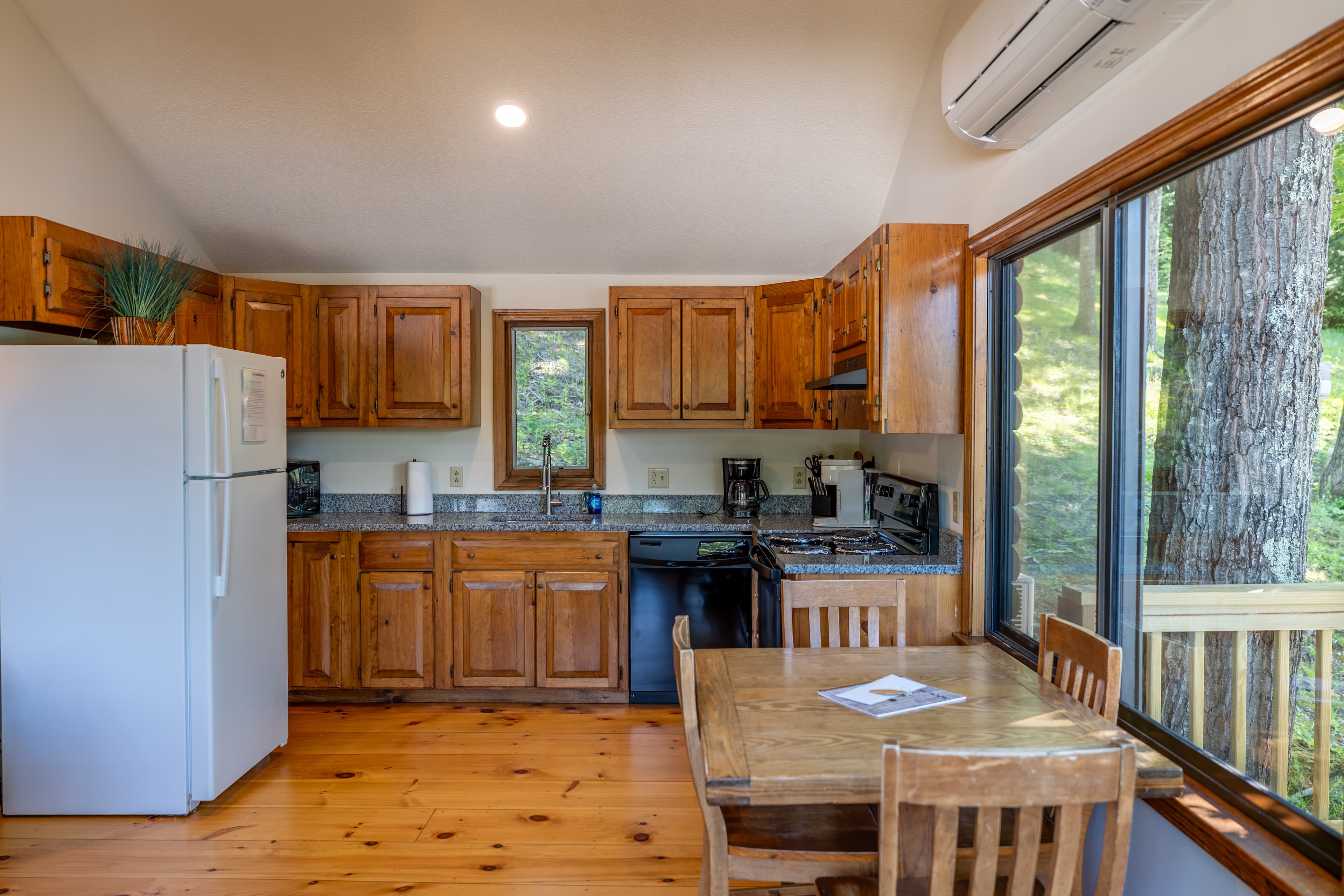 A bright cabin kitchen and dining area with a vaulted ceiling, hardwood floors, wood cabinets, a white fridge, and a wooden dining set.