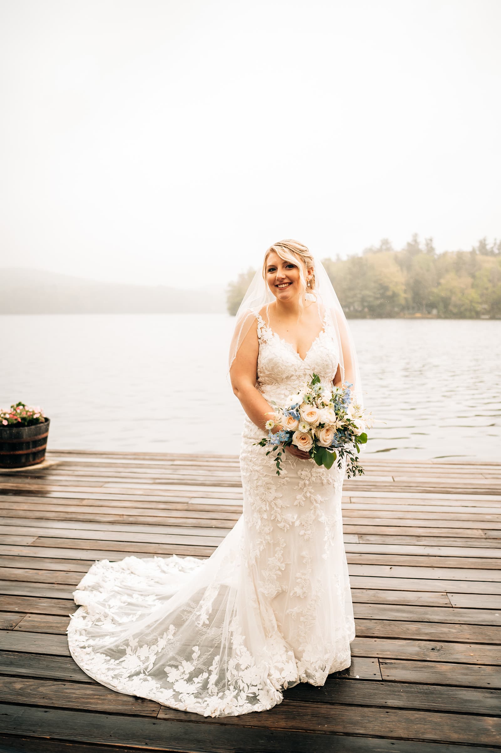 A smiling bride in a lace wedding dress stands on a wooden dock by a serene lake, holding a bouquet of flowers.
