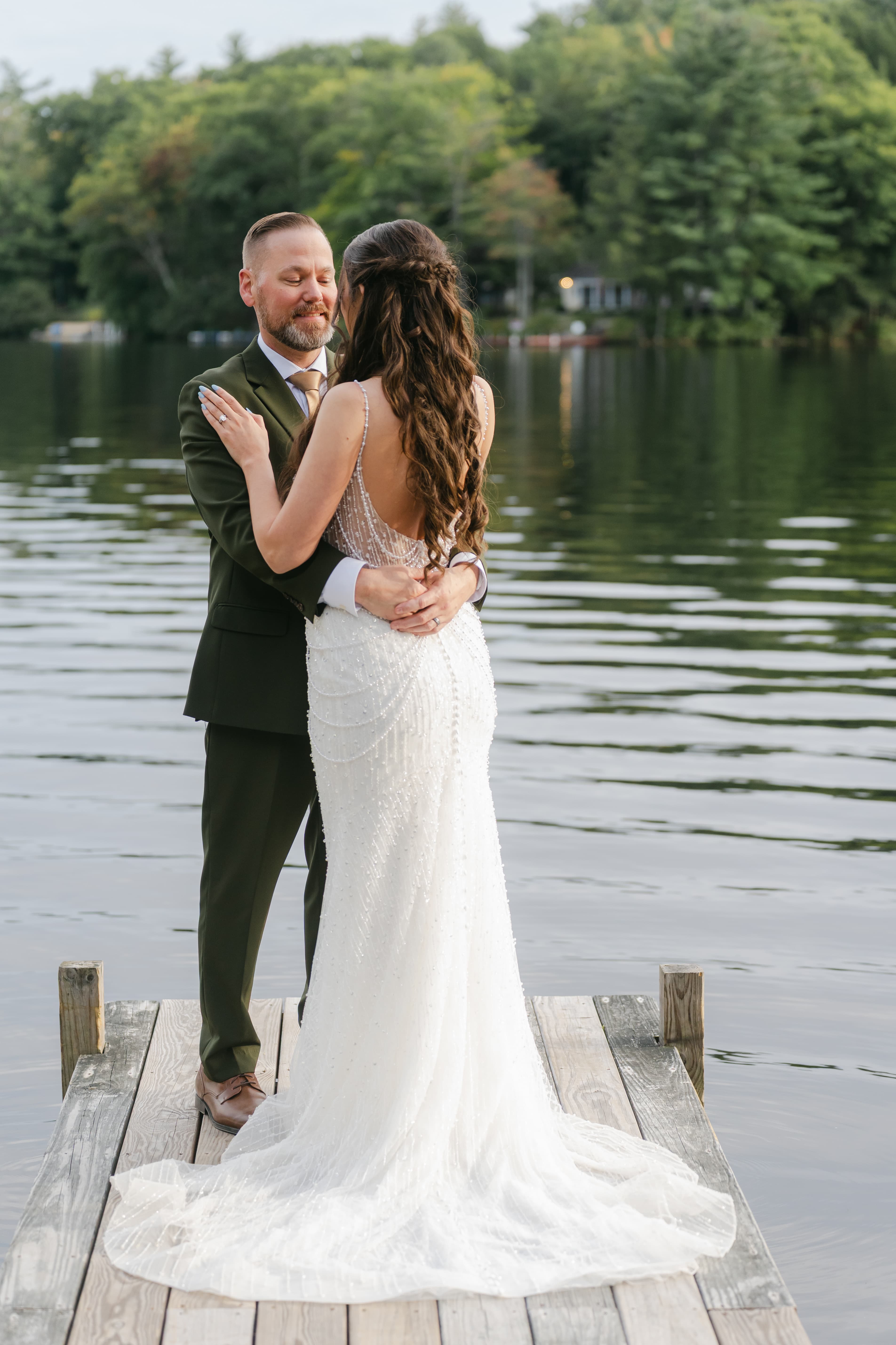 A couple in wedding attire embraces on a dock by a serene lake.