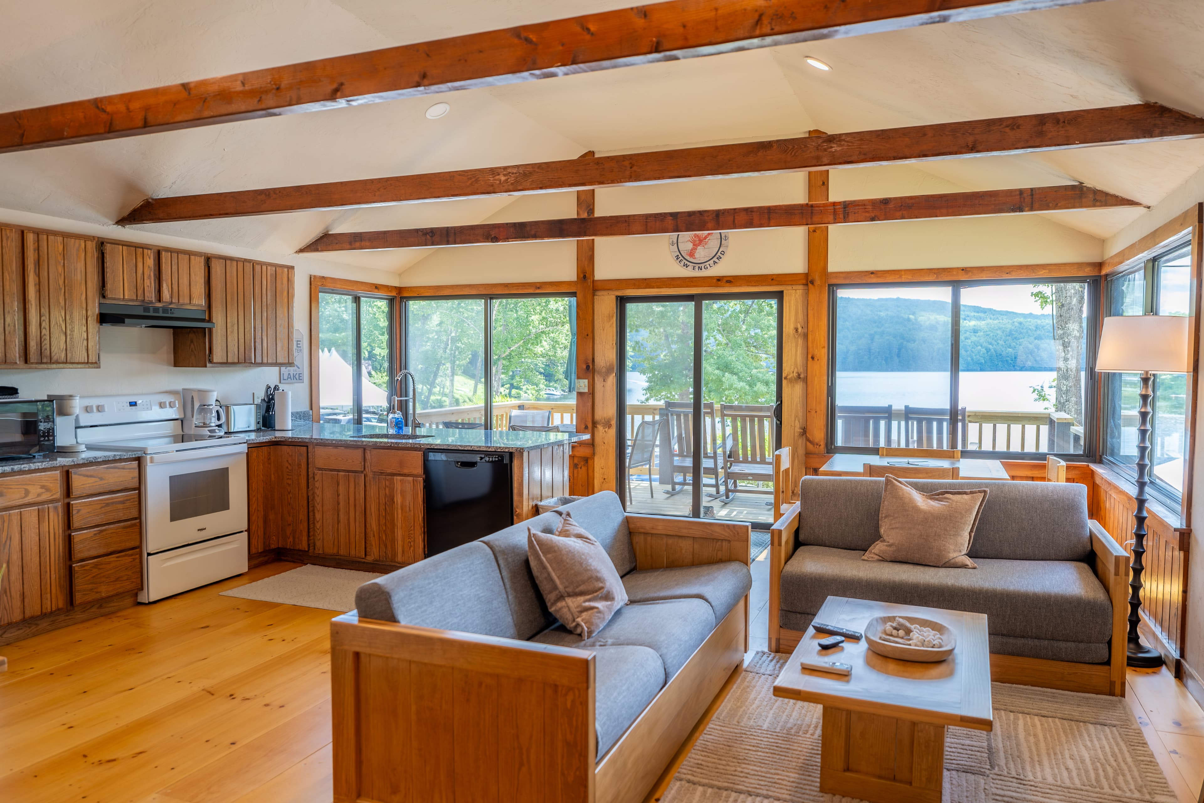 An open-plan cabin interior with exposed wooden beams, featuring a kitchen, dining area, and living space opening onto a deck with lake views.