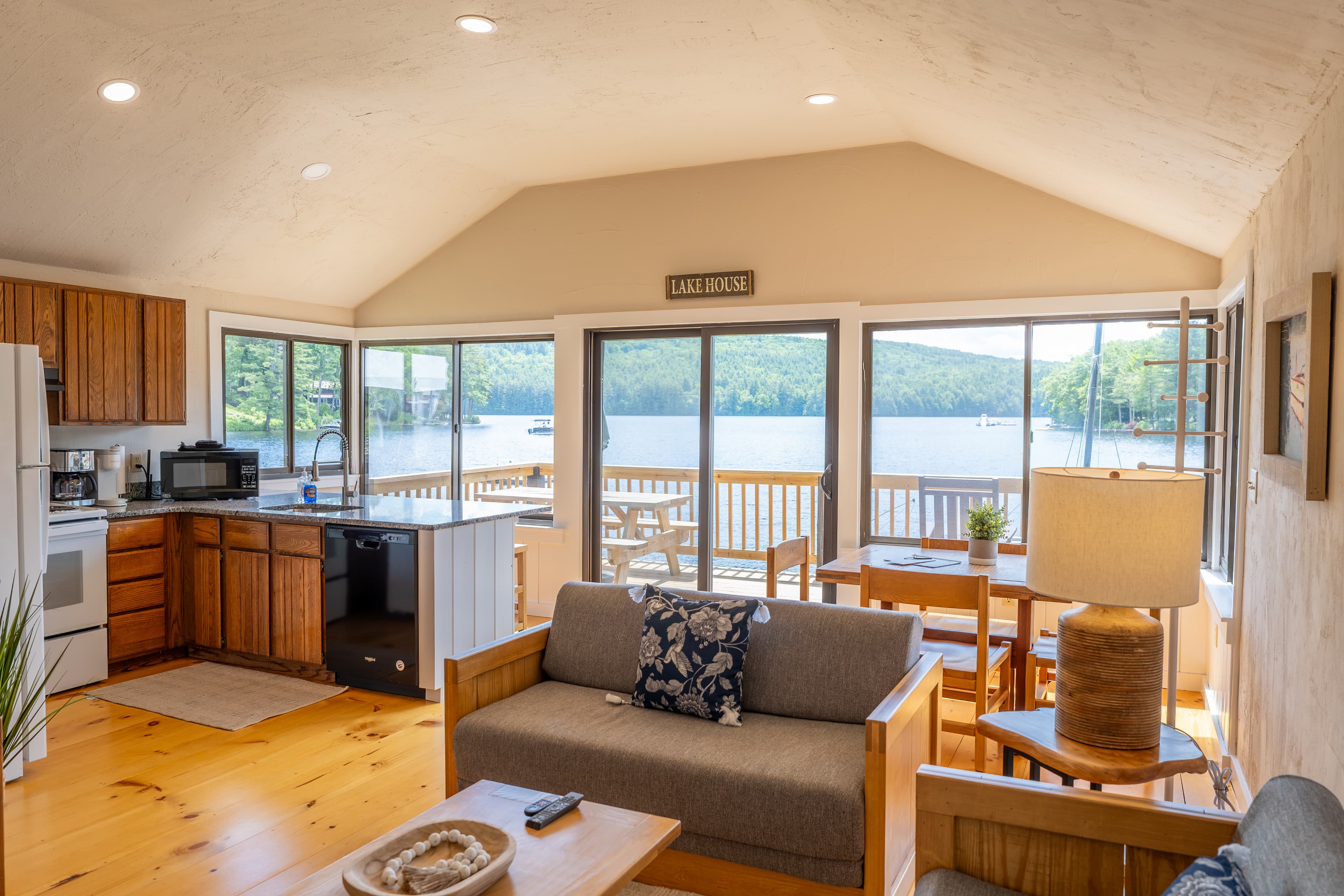 An open-concept cabin space with a vaulted ceiling, featuring a living area, kitchen with wood cabinets, and large windows overlooking a lake.