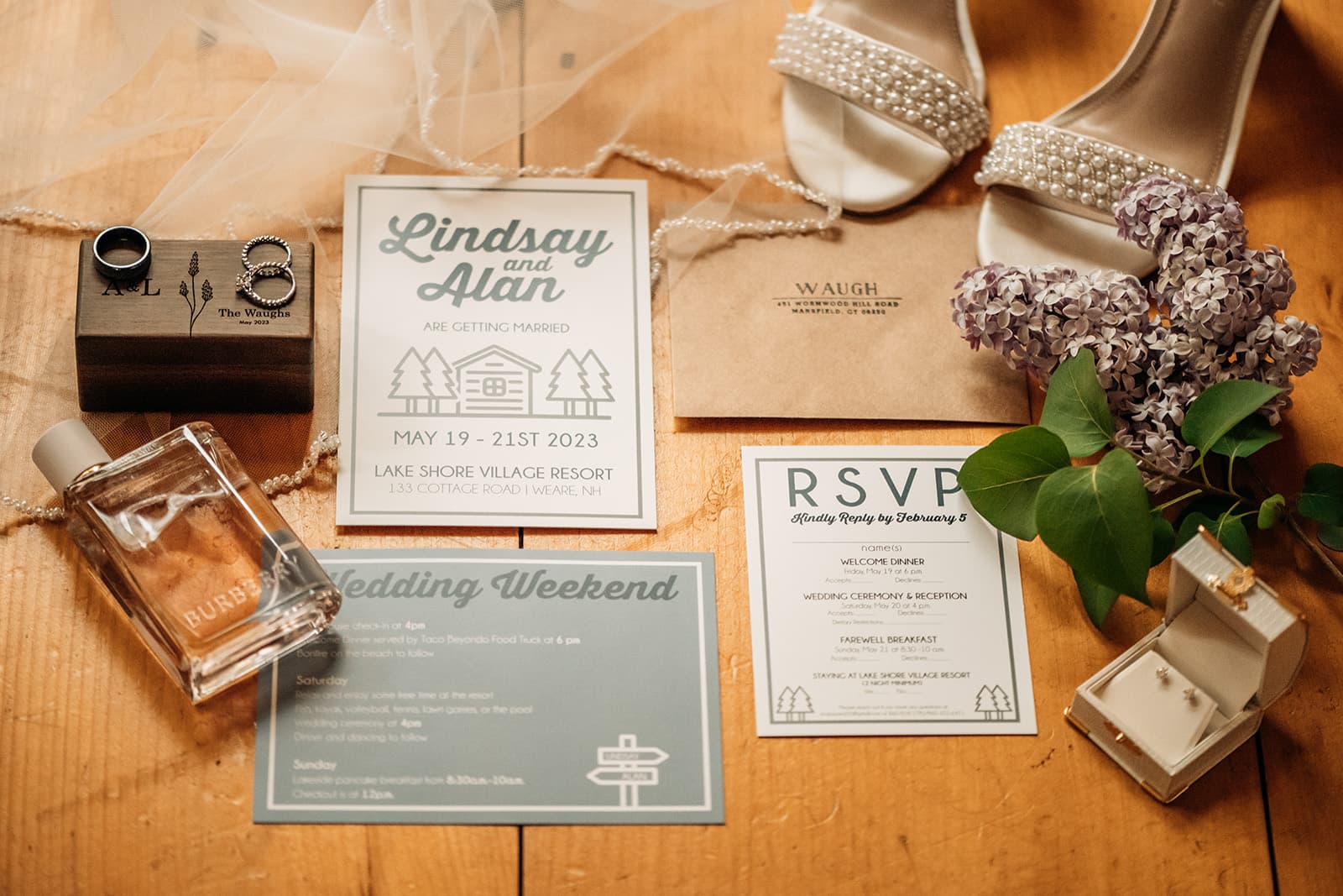 Flat lay of wedding invitation materials, including rings, perfume, RSVP, and a bouquet of lilacs.
