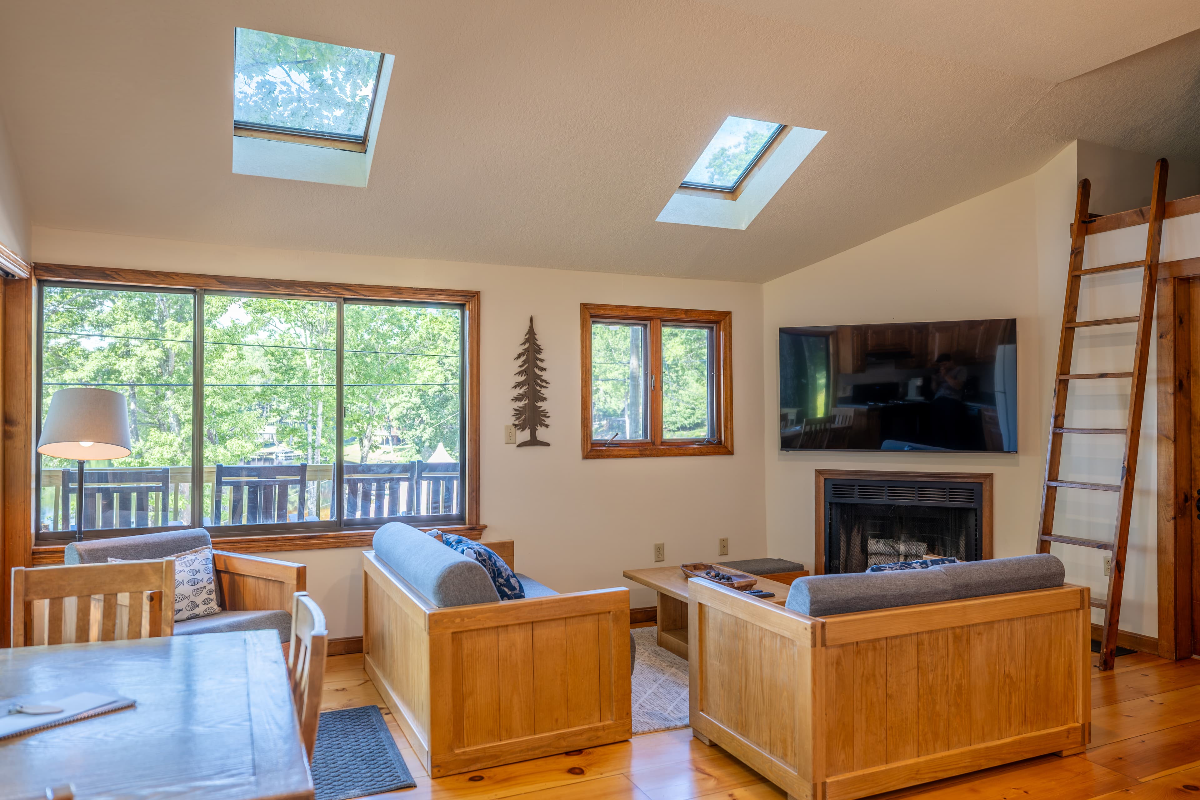 A bright cabin living room with a skylight, hardwood floors, a blue futon, a small fireplace, and a ladder leading to a loft area.