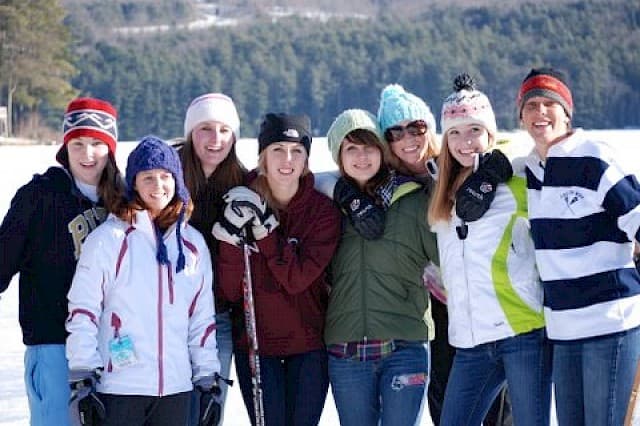 A group of eight young women stands together, smiling on a snowy landscape, wearing winter clothing and hats.
