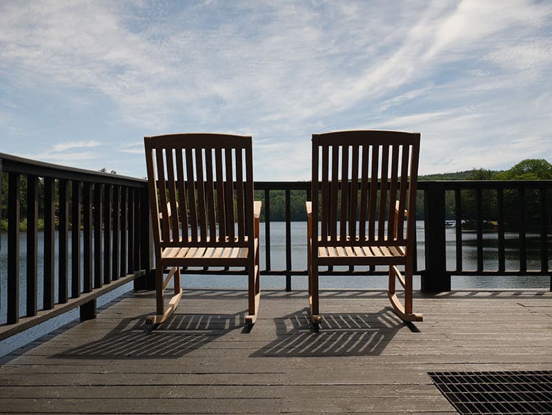 Two wooden rocking chairs face a calm lake under a blue sky.