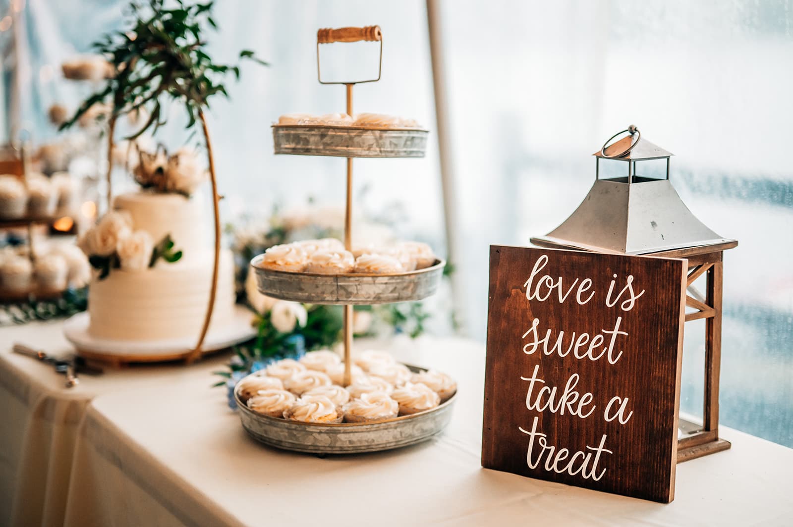 A dessert table featuring a tiered tray of cupcakes, a wedding cake, floral decorations, and a sign that says "love is sweet take a treat."