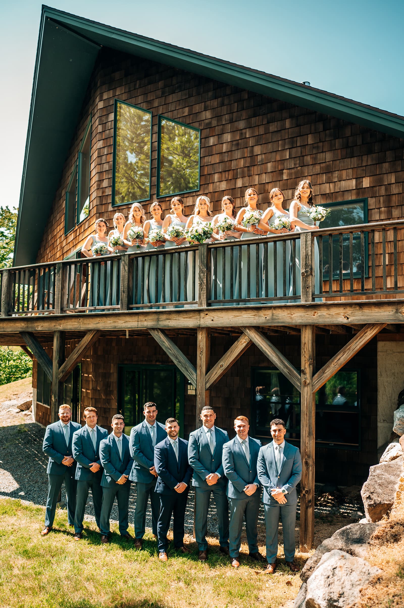 A bridal party in elegant attire poses outside a rustic wooden house, with bridesmaids on the balcony and groomsmen standing below.