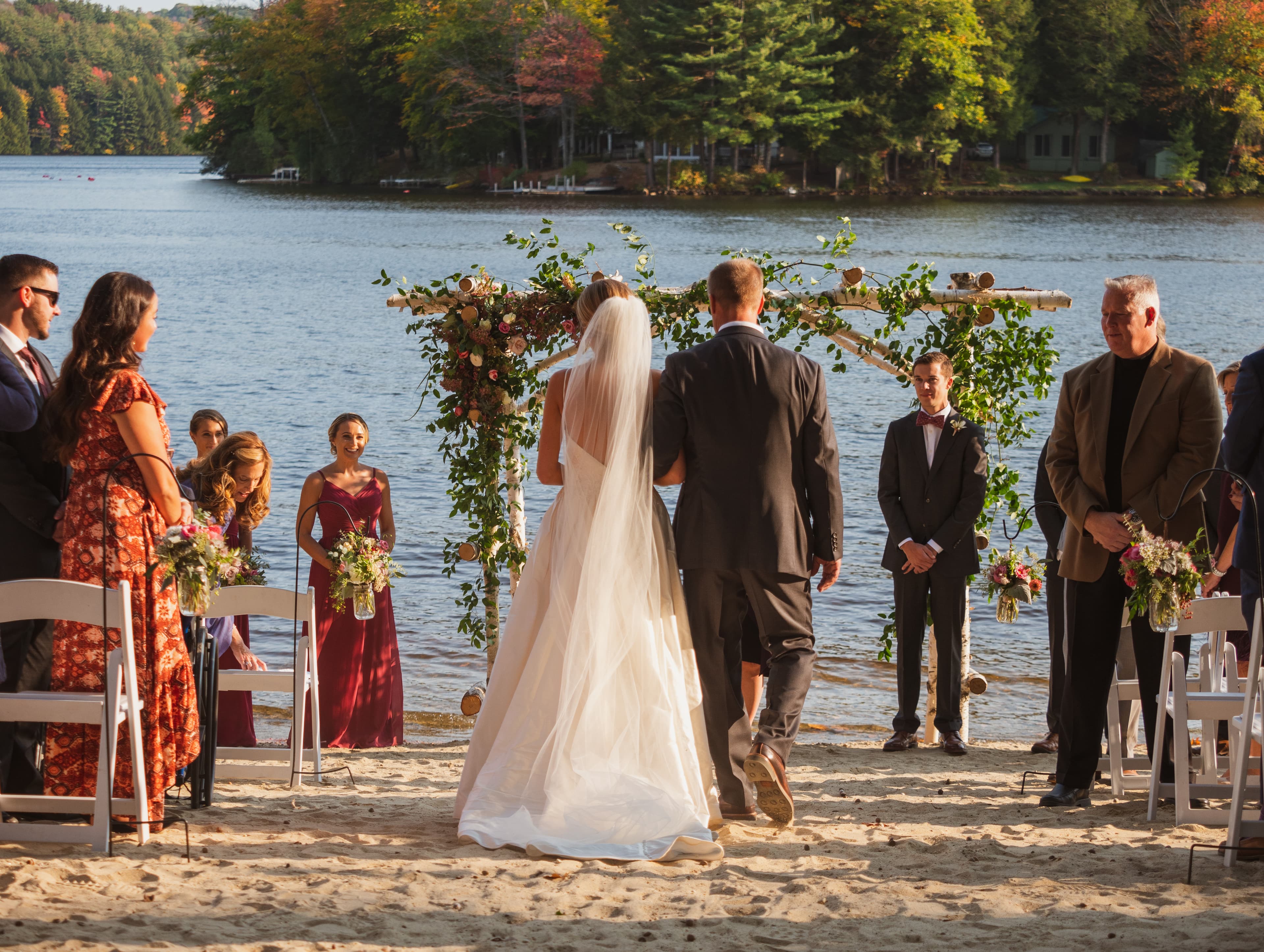 A bride and groom stand at the altar by a lake, surrounded by wedding guests in a picturesque autumn setting.