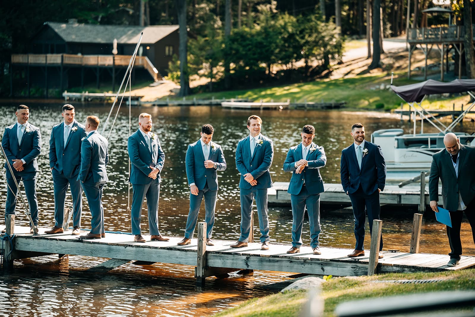 A group of seven men in suits stands on a dock by a lake, with trees and a house in the background.