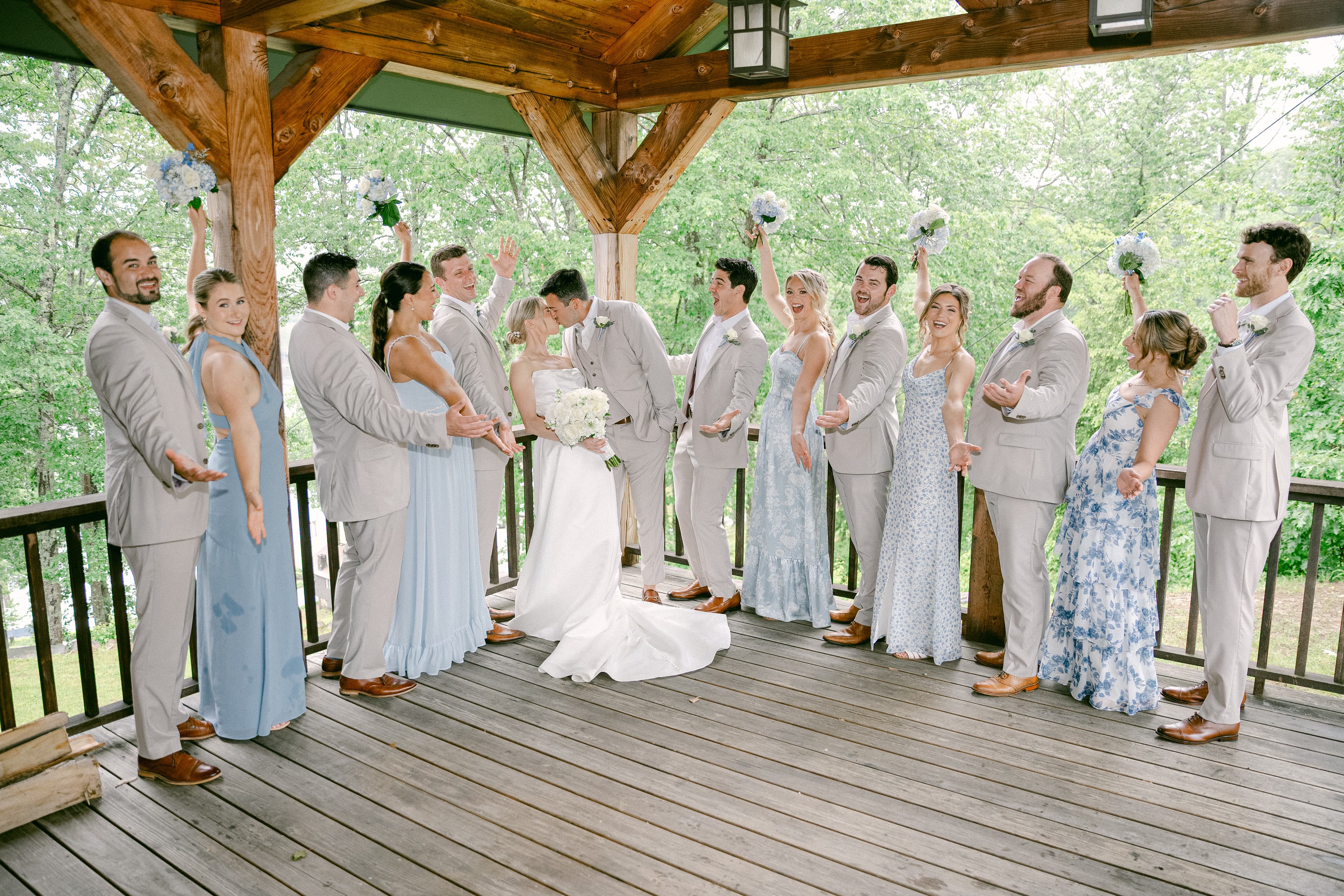 A bride and groom kiss at their wedding surrounded by their bridal party on a wooden deck.