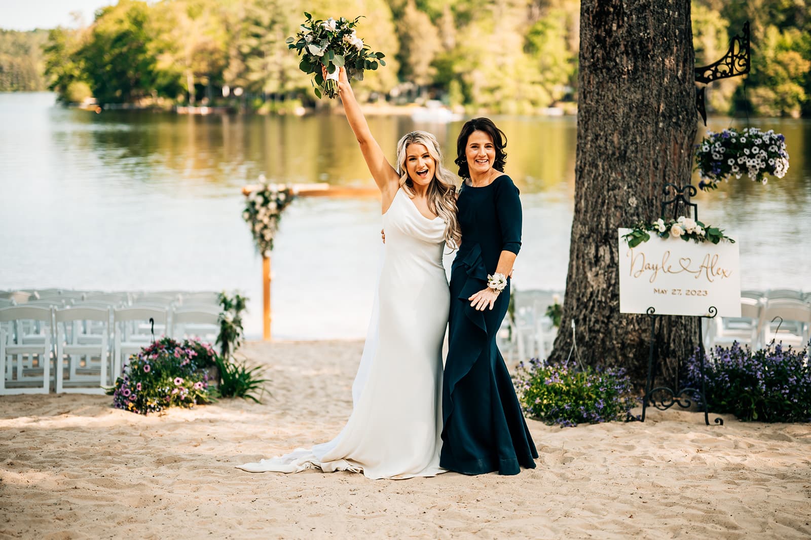 A joyful bride in a white gown raises her bouquet alongside a woman in a dark dress at a lakeside wedding.