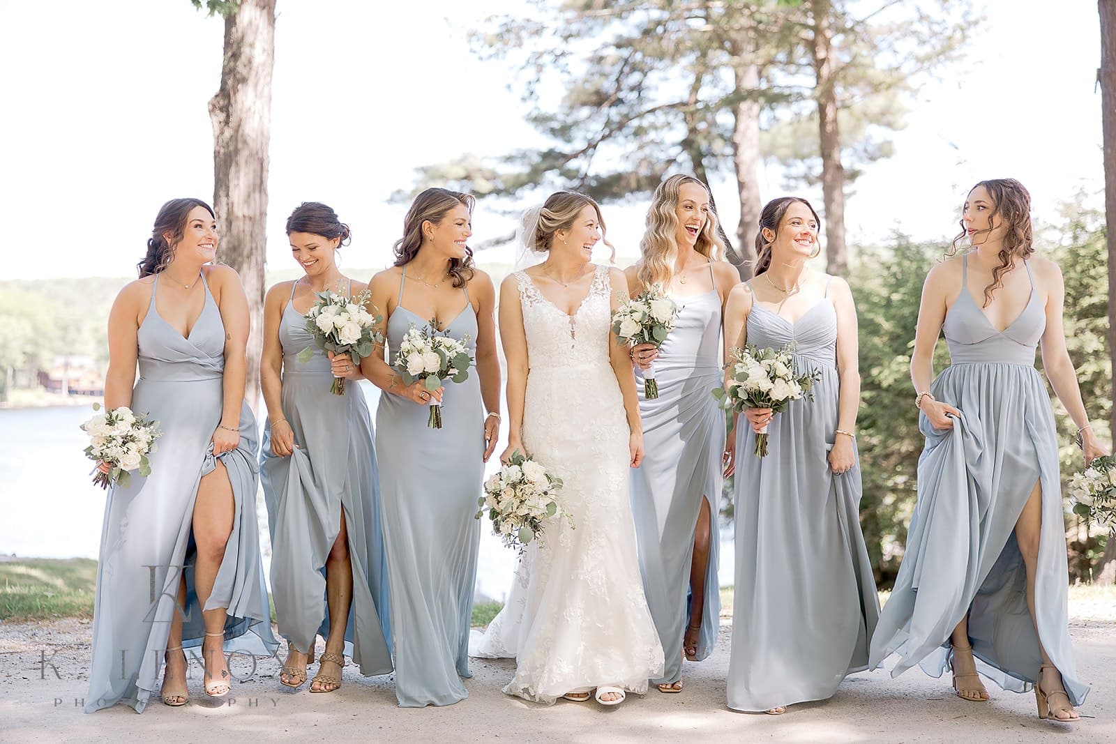 A bride in a white gown walks with her bridesmaids, who are wearing light blue dresses, surrounded by trees and a lake.