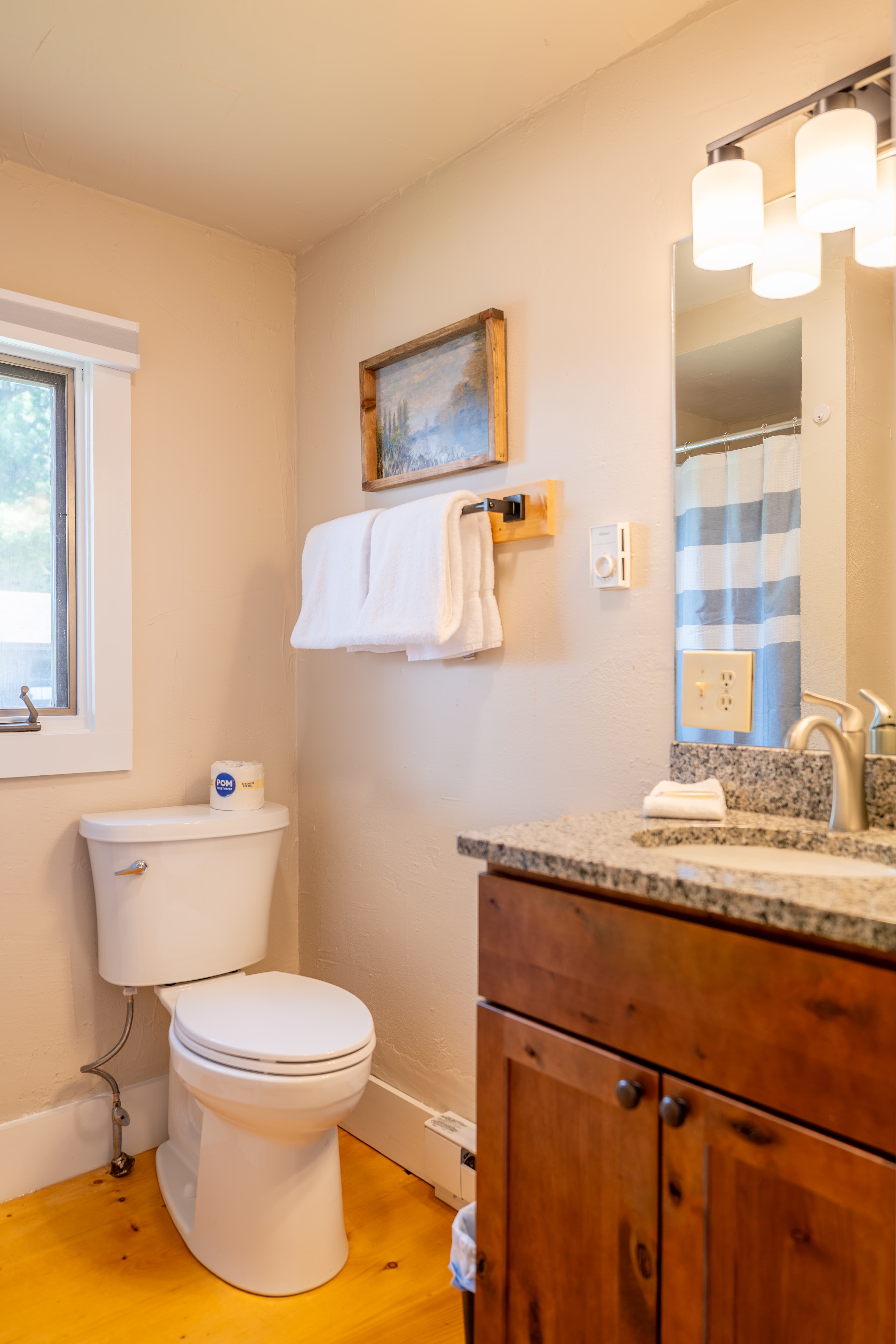 A simple, well-lit bathroom with a white toilet, wooden vanity, towels on a wooden rack, and a small, curtained window.