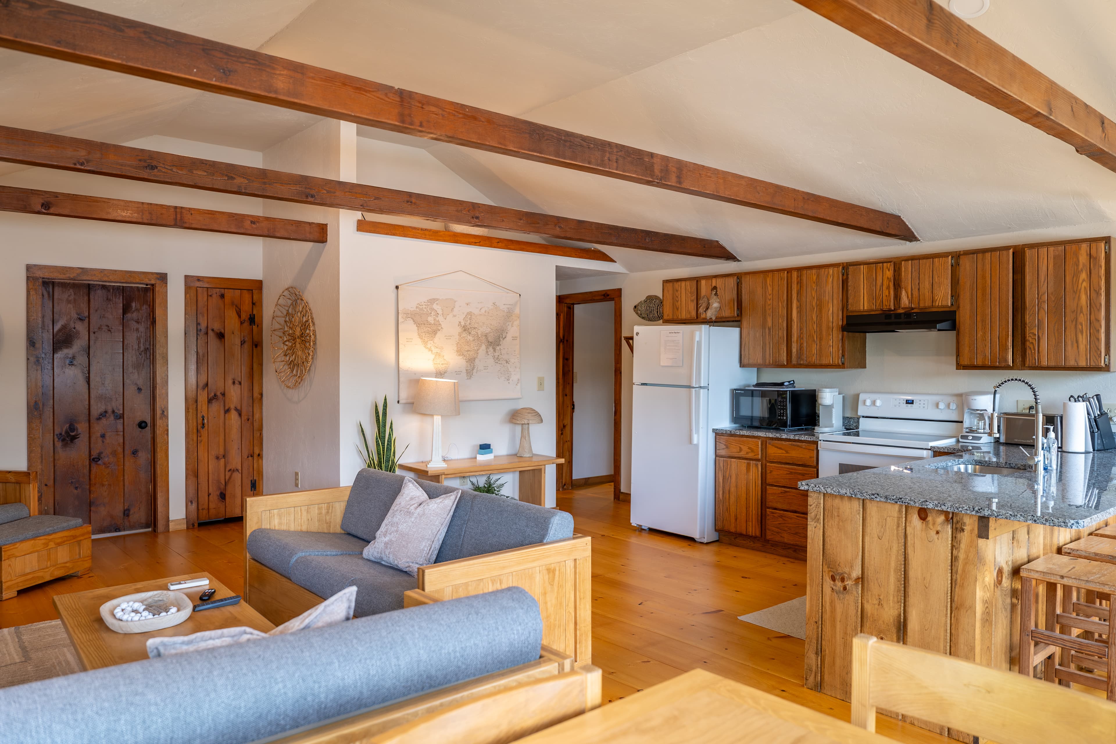 A cozy living space featuring exposed wooden beams, modern globe lights, a sofa, armchair, and a wicker dining set.