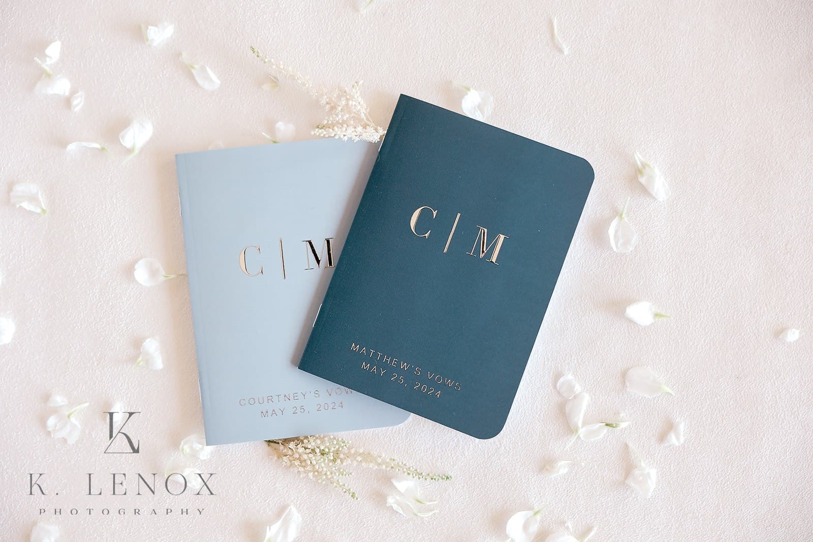 Two vow booklets with gold lettering on a light background, surrounded by scattered flower petals.