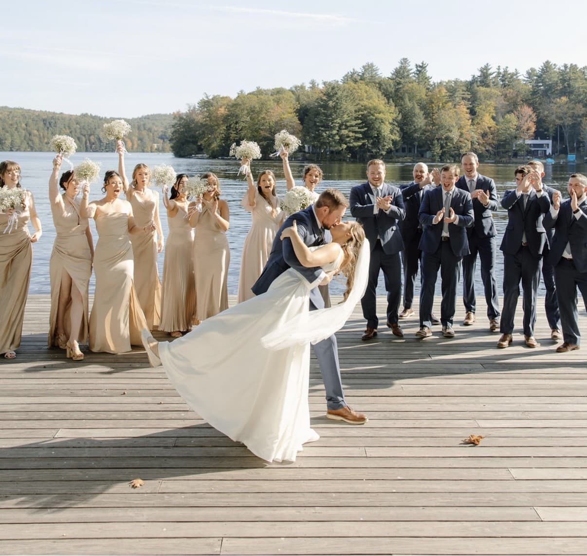 A couple shares a kiss on a dock while their wedding party celebrates in the background.