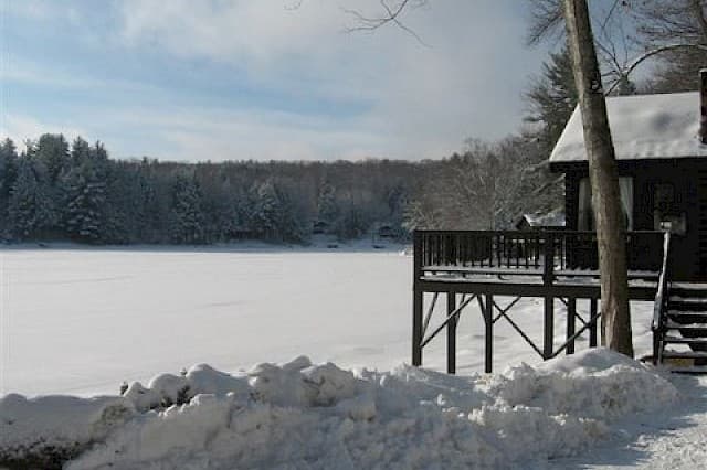 A snowy lakeside scene with a wooden deck and evergreen trees in the background.