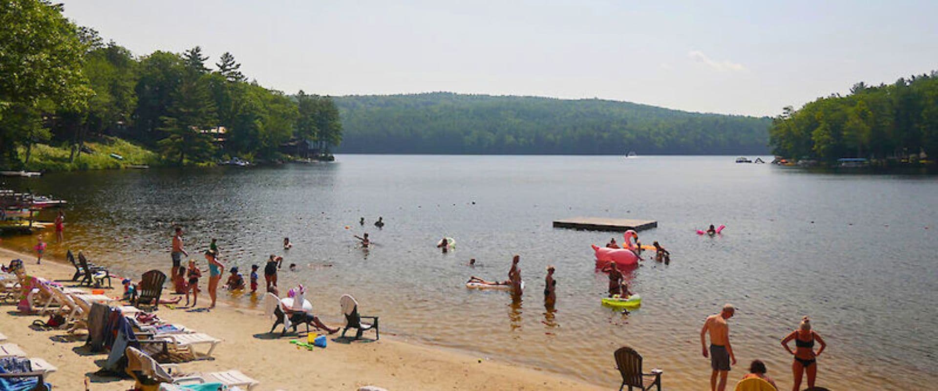 A sandy beach by a lake filled with people swimming and sunbathing on a sunny day.